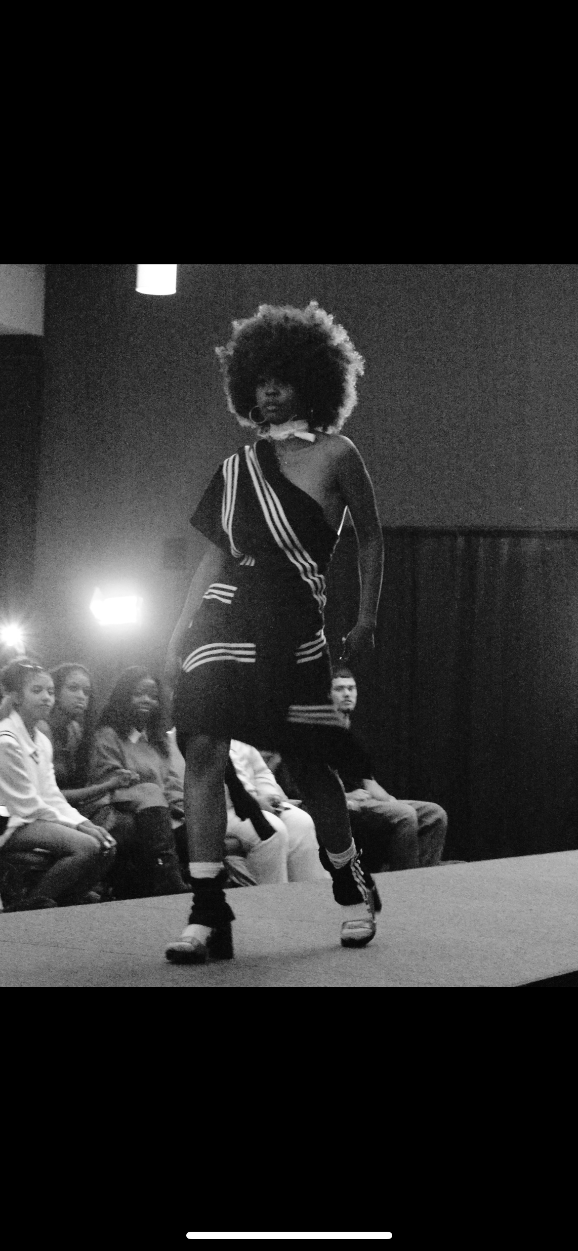 A woman walking on a runway with an audience watching, wearing a stylish black dress with white stripes, high heels, and sporting curly hair.