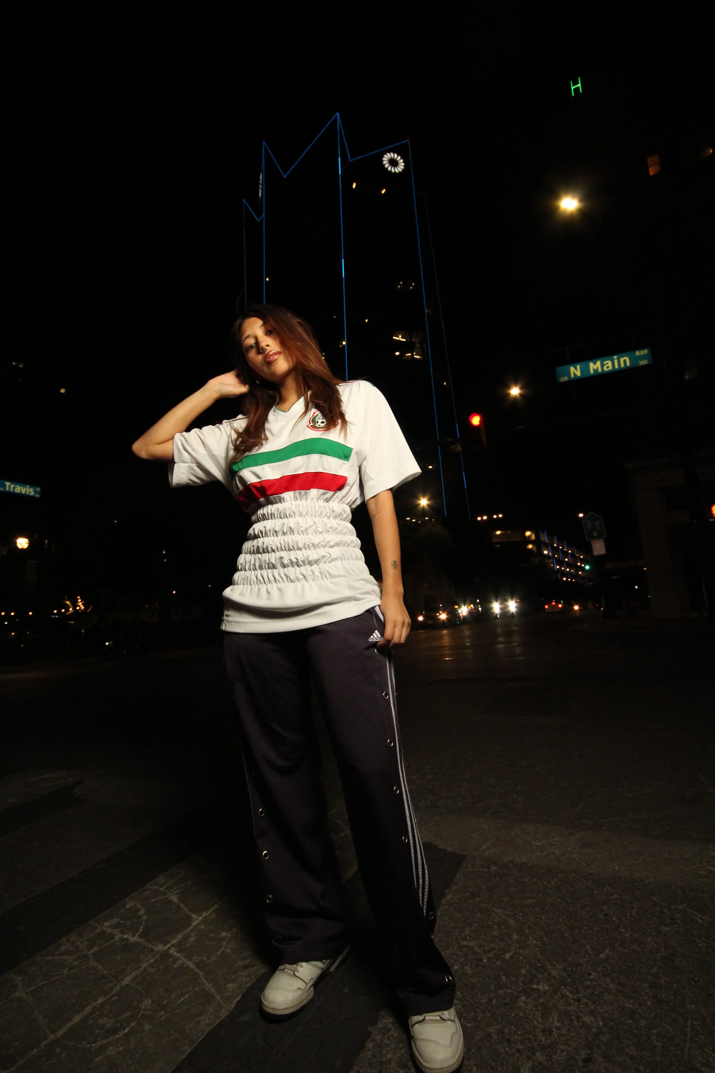 A woman standing on a city street at night, wearing a sports jersey, black pants, and sneakers, with streetlights and tall buildings in the background.
