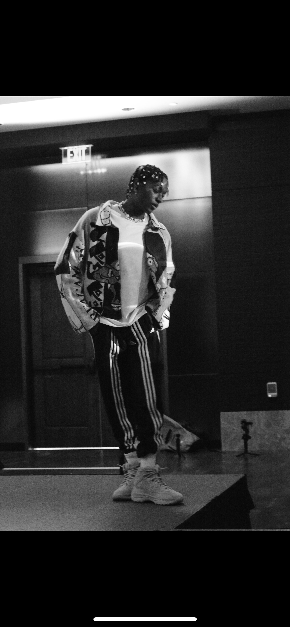 A person with braided hair stands on a stage, wearing a patterned jacket, a white shirt, and Adidas track pants, inside a room with dark walls and an exit sign in the background.