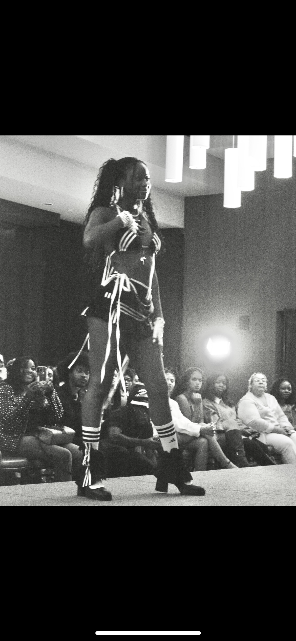 A woman walking on a runway at a fashion show, wearing athletic-inspired clothing with stripes and high-heeled shoes, audience members watching attentively.