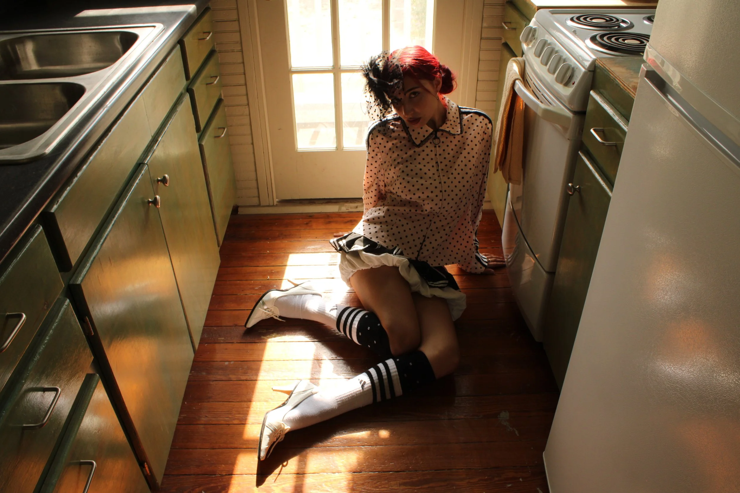 A woman with red hair and wearing a polka-dot shirt is kneeling on a kitchen floor, leaning against a cabinet, with her head tilted downward. The kitchen has wooden flooring, green cabinets, and white appliances. Sunlight is coming through a glass do