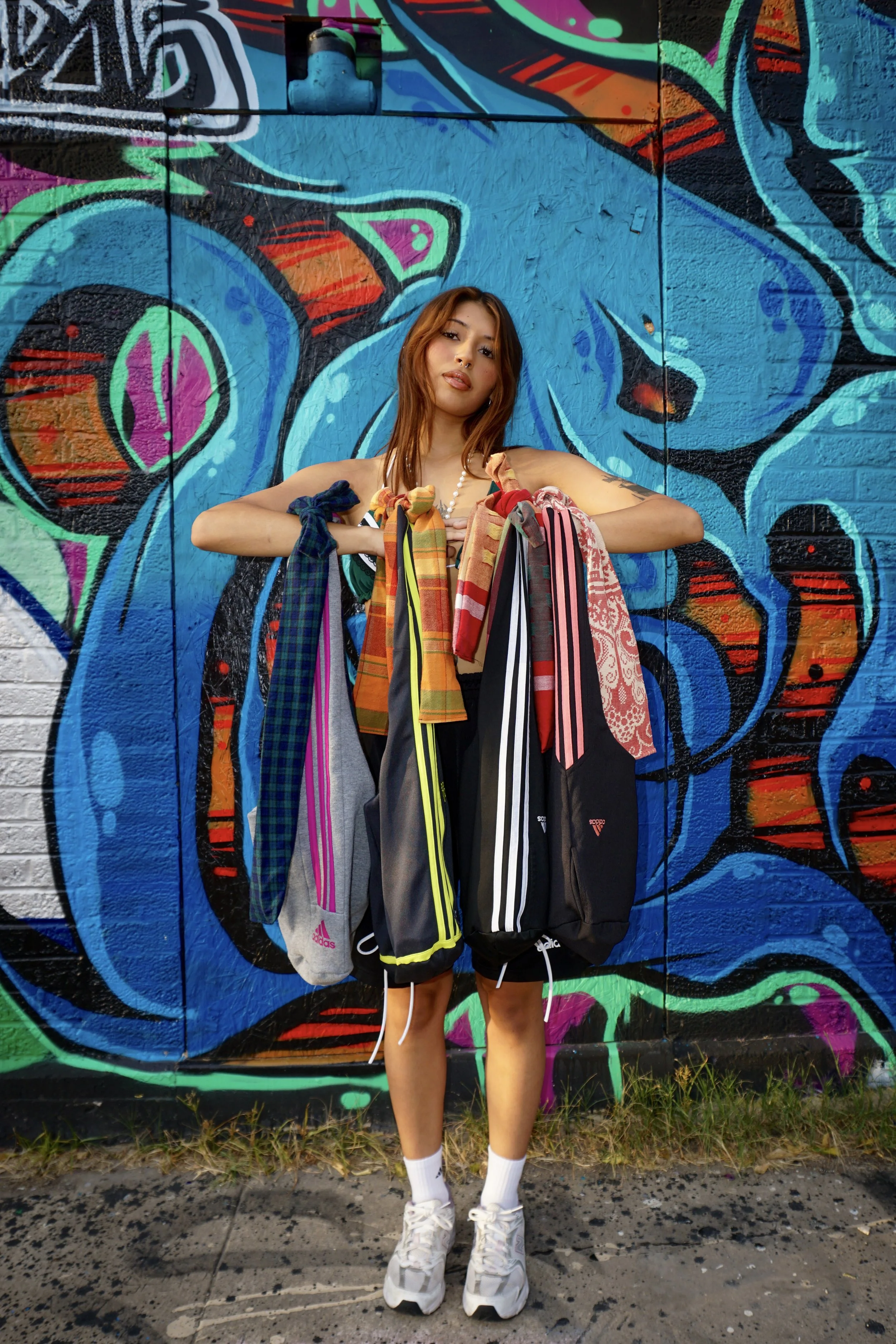 A young woman standing in front of a colorful graffiti wall, holding multiple Adidas clothing items with striped and patterned designs.