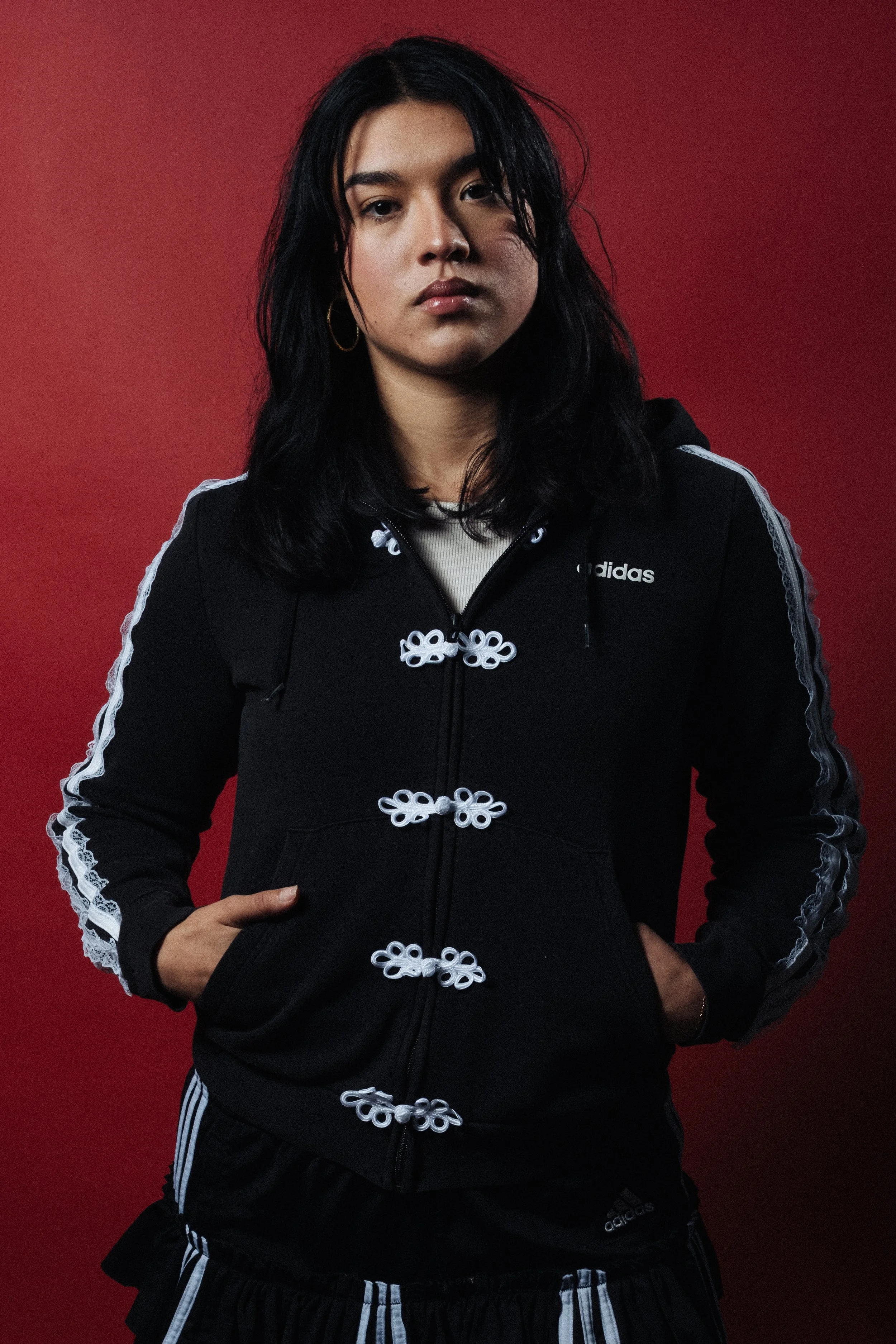 A woman with black hair wearing a black Adidas hoodie with white striped details on the sleeves, standing against a red background.