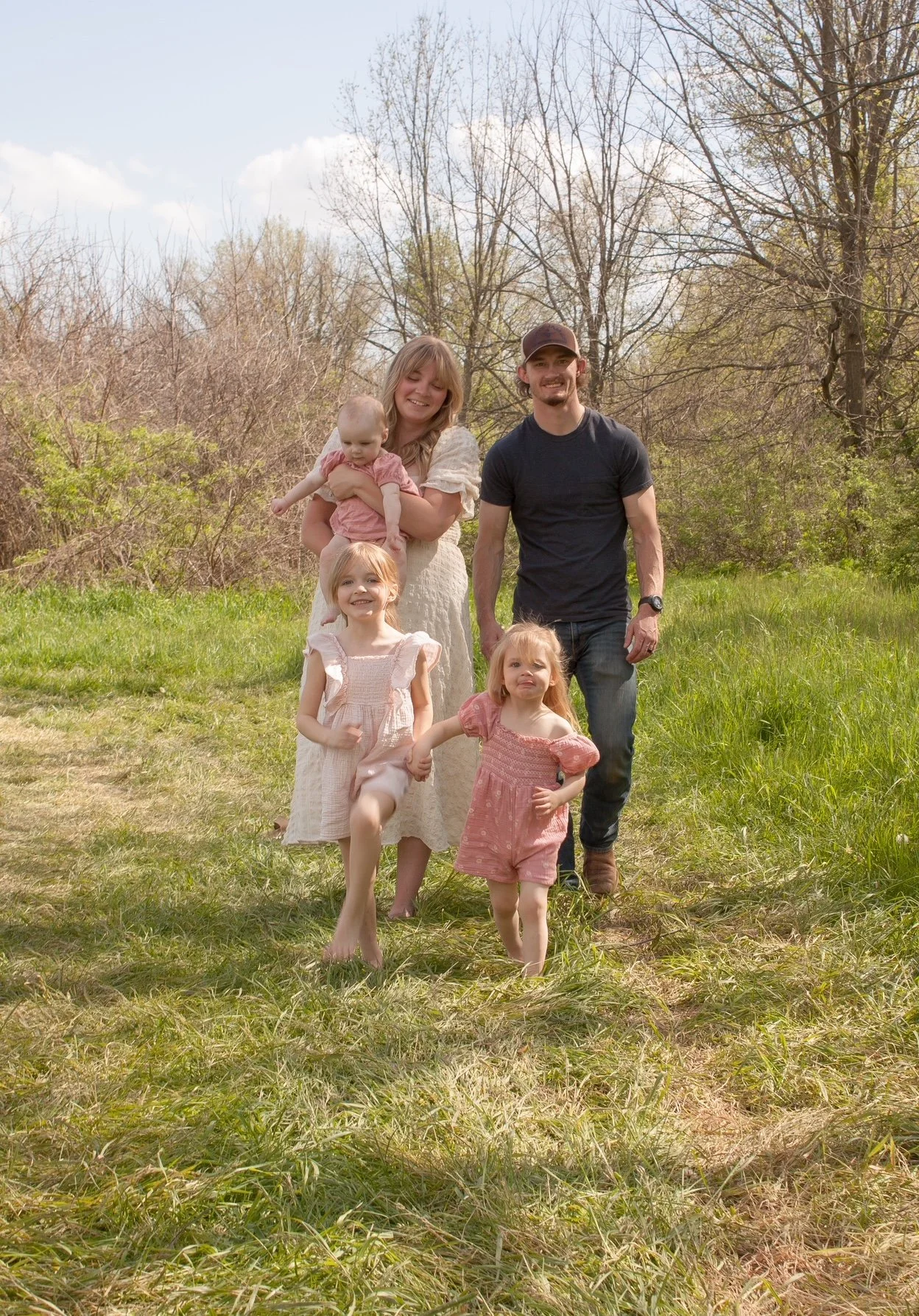 A family of six walking outdoors on a grassy field with trees and a cloudy sky in the background. The mother is holding a baby, and there are three young girls, two of them holding hands, standing in front of her. The father is walking beside them, smiling.