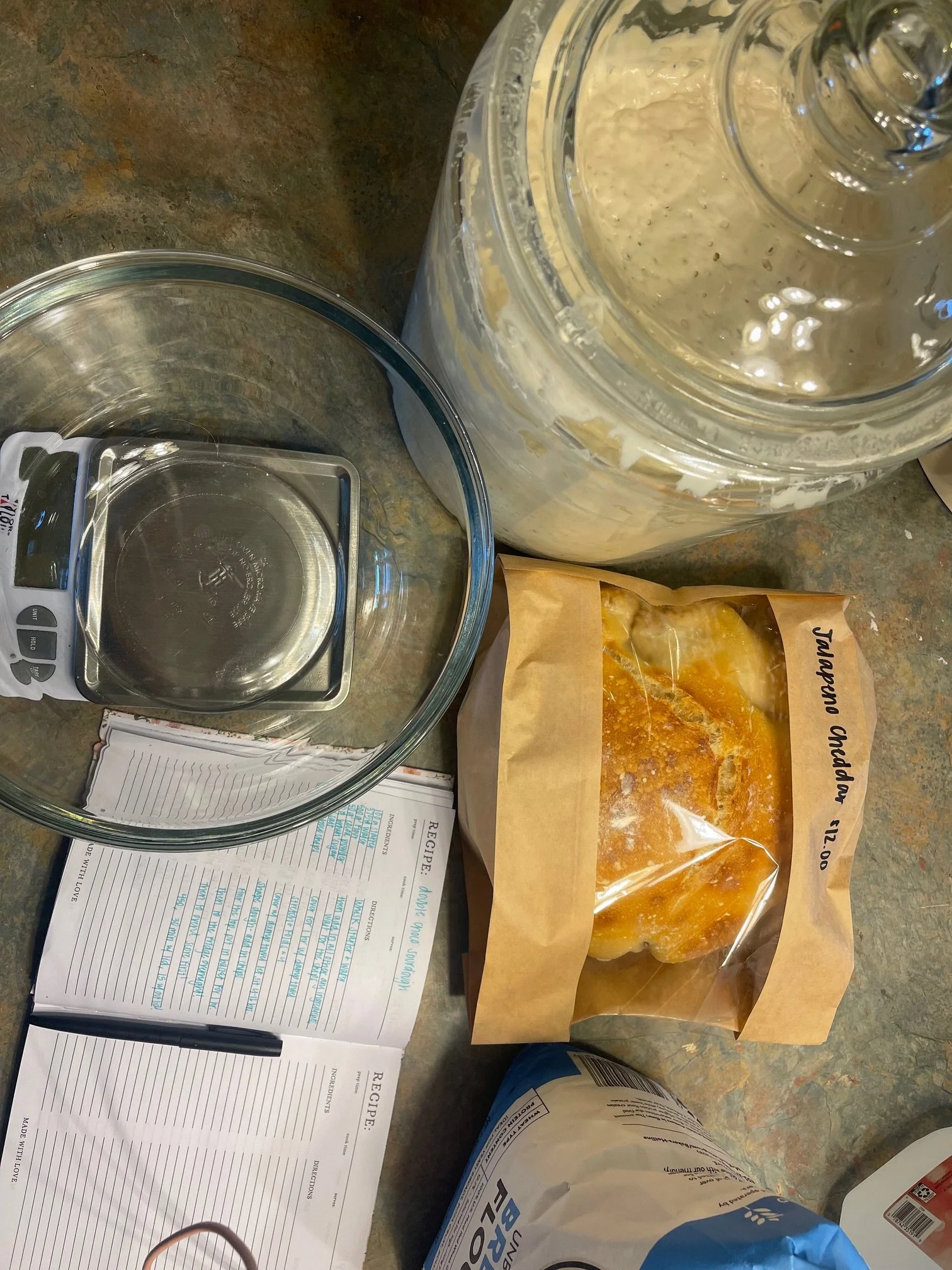 A kitchen counter with a large container of whipped mixture, a digital scale, a bag of flour, a recipe notebook, and a packaged sourdough loaf.