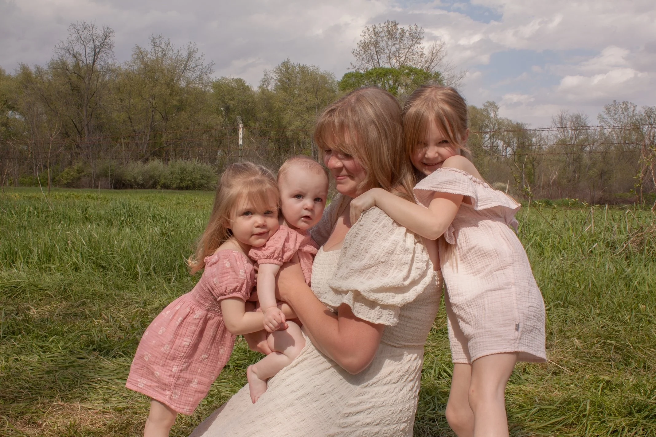 A woman with long hair holding four young girls outdoors in a grassy field with trees and cloudy sky in the background.