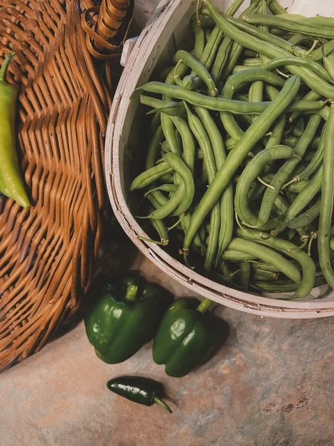 Fresh green beans in a basket, with two green bell peppers and a jalapeño on the floor in front of the basket.
