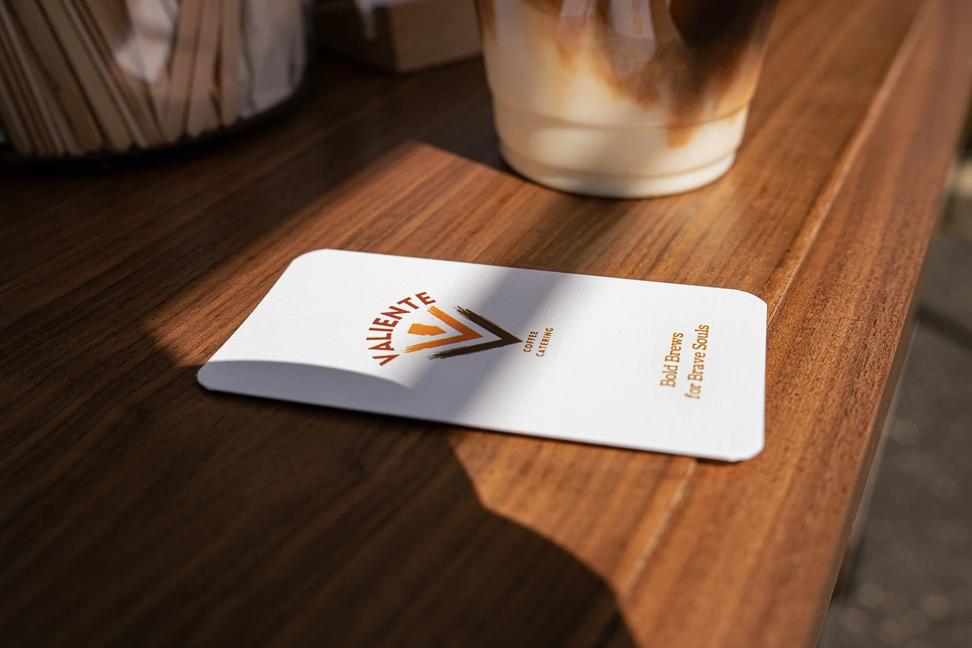 Close-up of a wooden table with a coffee cup and a menu from Valiente Coffee. The business card has the words 'Bold Brews for Brave Souls' printed on it.