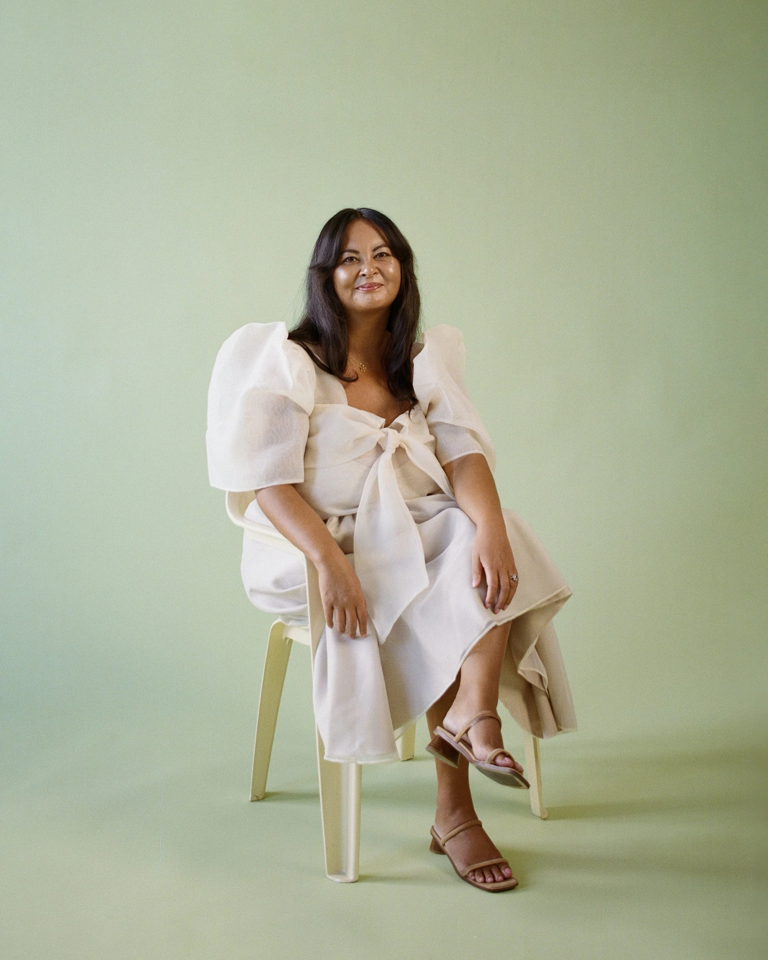 A woman sitting on a yellow chair against a light green background. She is wearing a Filipiniana top with a bow at the front, and tan sandals. She is smiling gently with her legs crossed at the ankle.