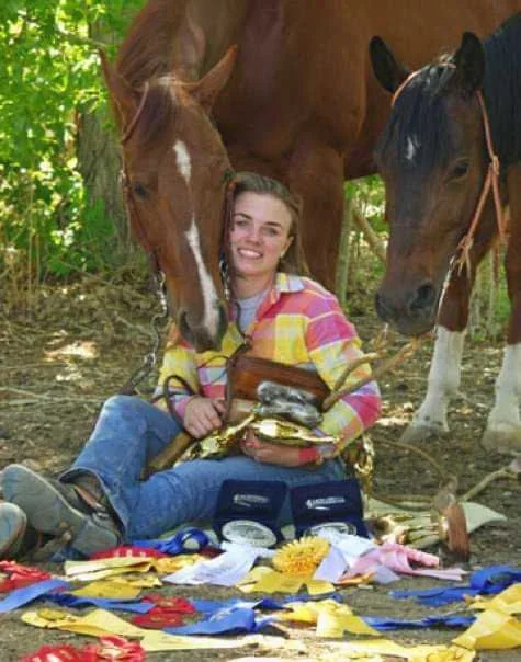 horse rider sitting on ground with an arm full of trophies and ribbons while two horses look over her shoulder