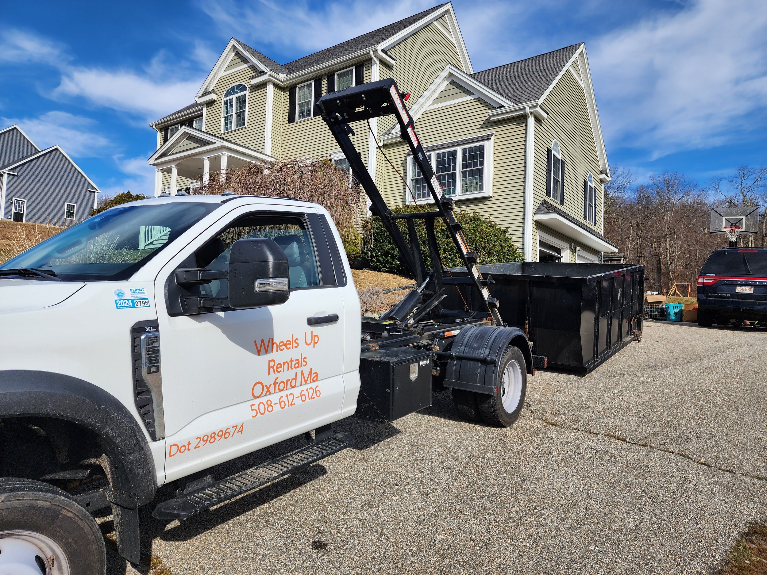 A white truck with a black bed and a lift, parked in front of a large yellow house with multiple stories, white trim, and multiple windows. There is a basketball hoop in the background and clear blue sky.