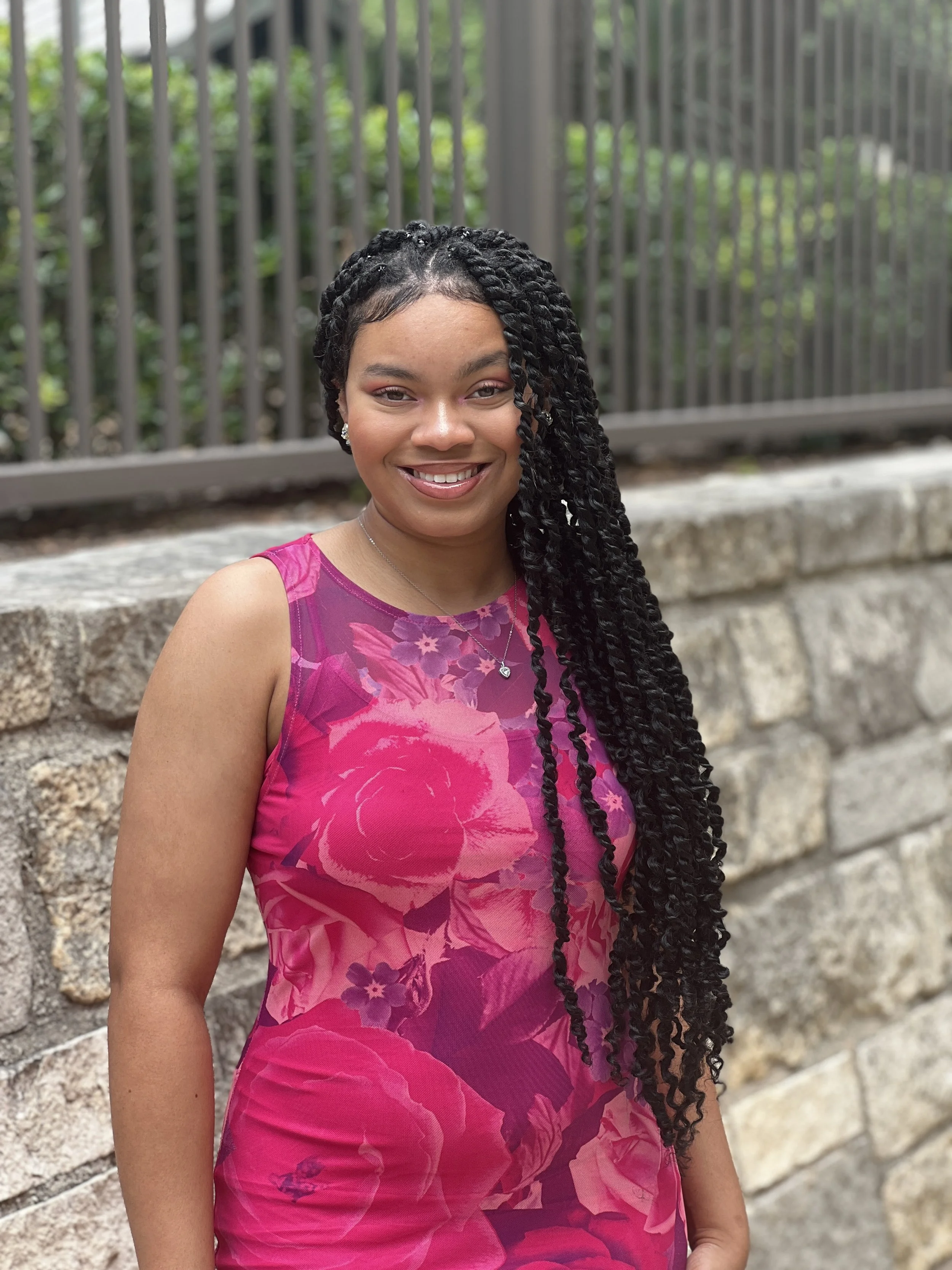 A woman with long black braided hair smiling outdoors in front of a stone wall with a black metal fence and green foliage in the background.