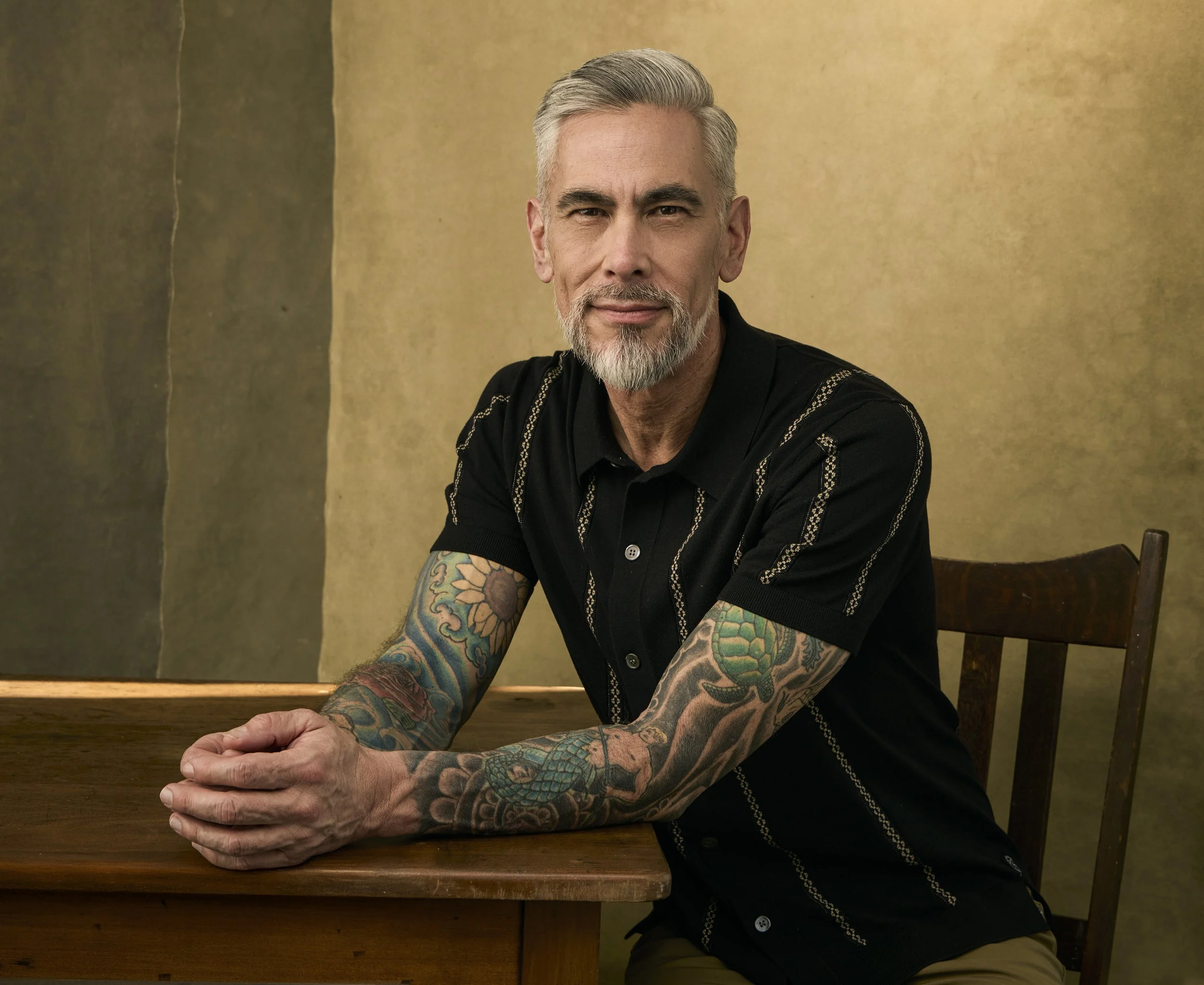 A middle-aged man with gray hair and a beard, wearing a black shirt with embroidered patterns and colorful tattoos on his arms, sitting at a wooden table with hands clasped, in front of a gold-colored background.