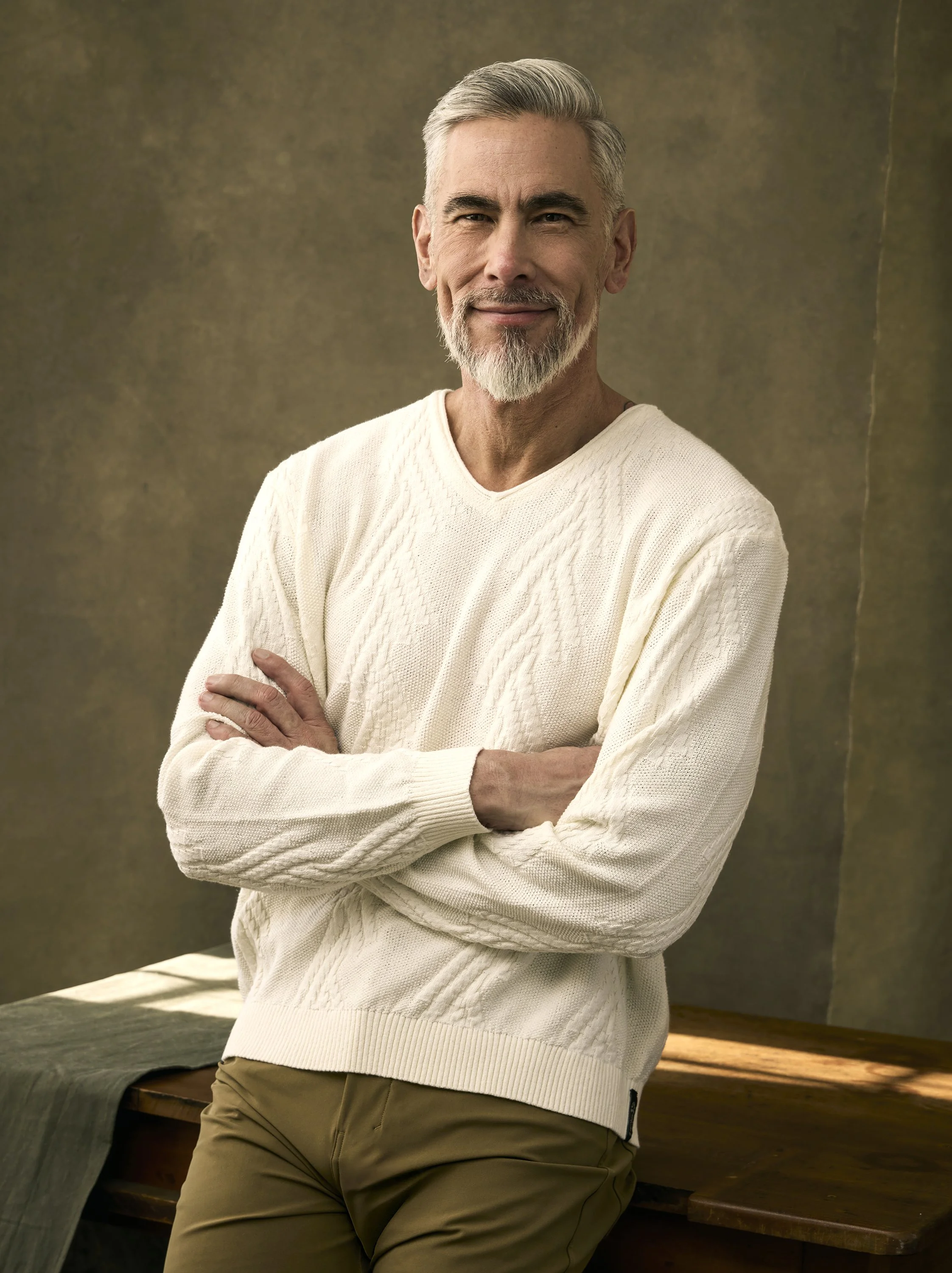 Close-up of a smiling older man with gray hair and beard, wearing a cream-colored sweater and khaki pants, standing with arms crossed in a room with a textured brownish-green wall background.