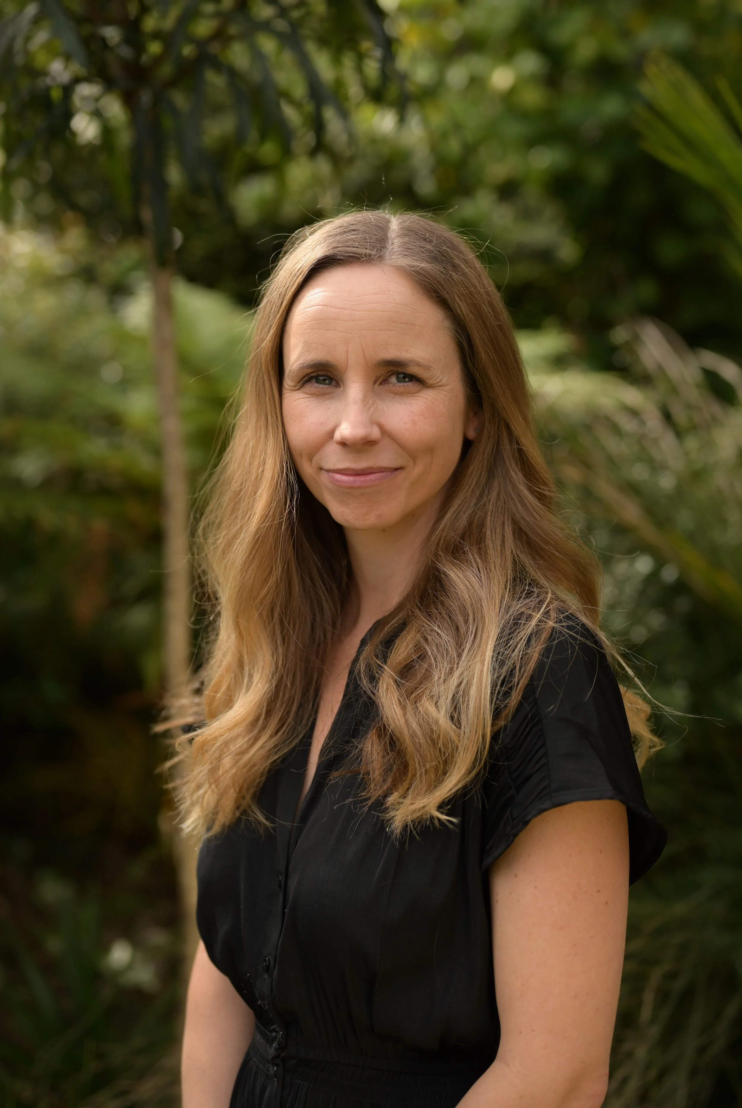 A woman with long, wavy blonde hair and light skin, smiling slightly, standing outdoors with green foliage and trees in the background.