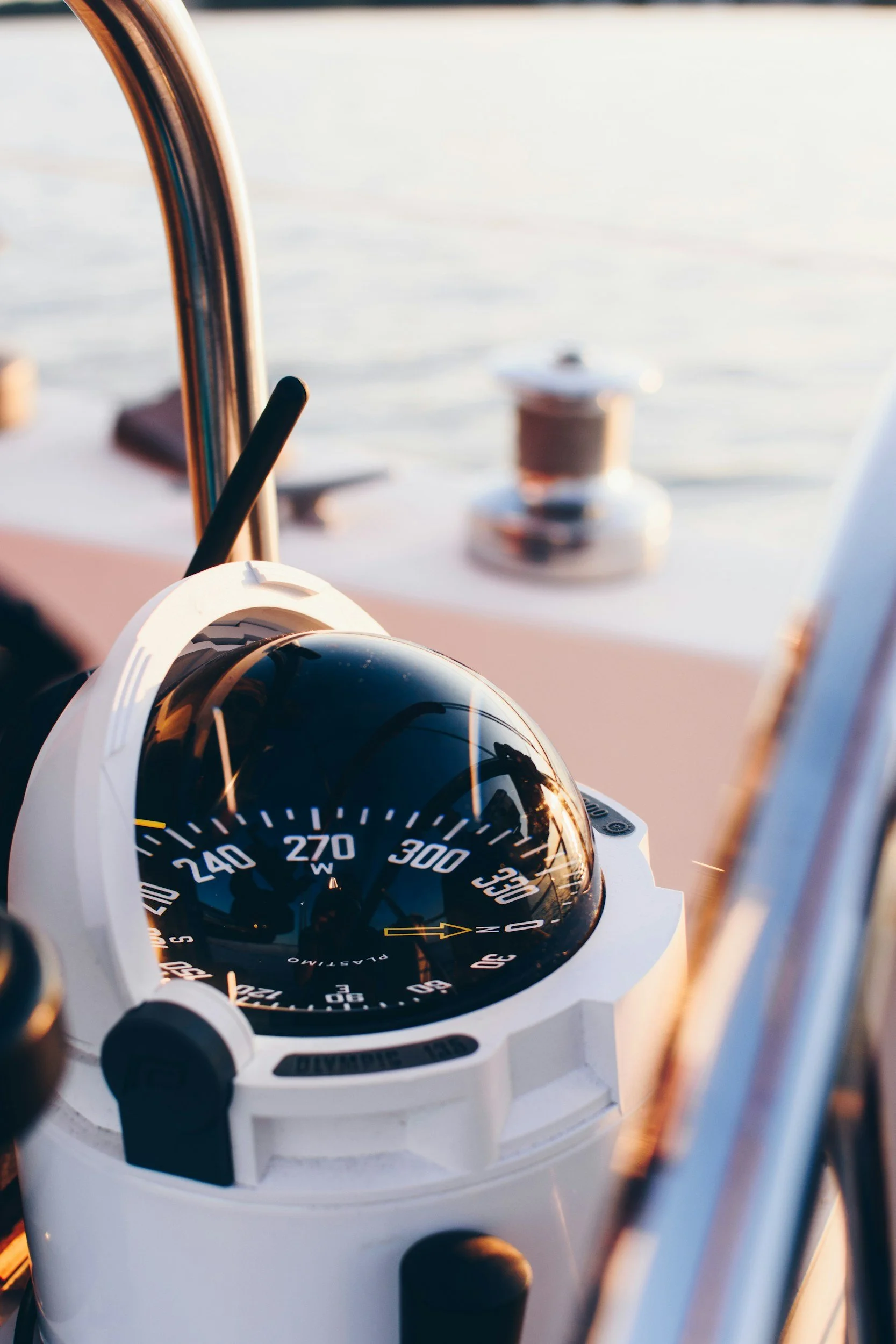 Close-up of a nautical compass mounted on a boat's dashboard, with a blurred view of the water and a mooring bollard in the background during sunset.