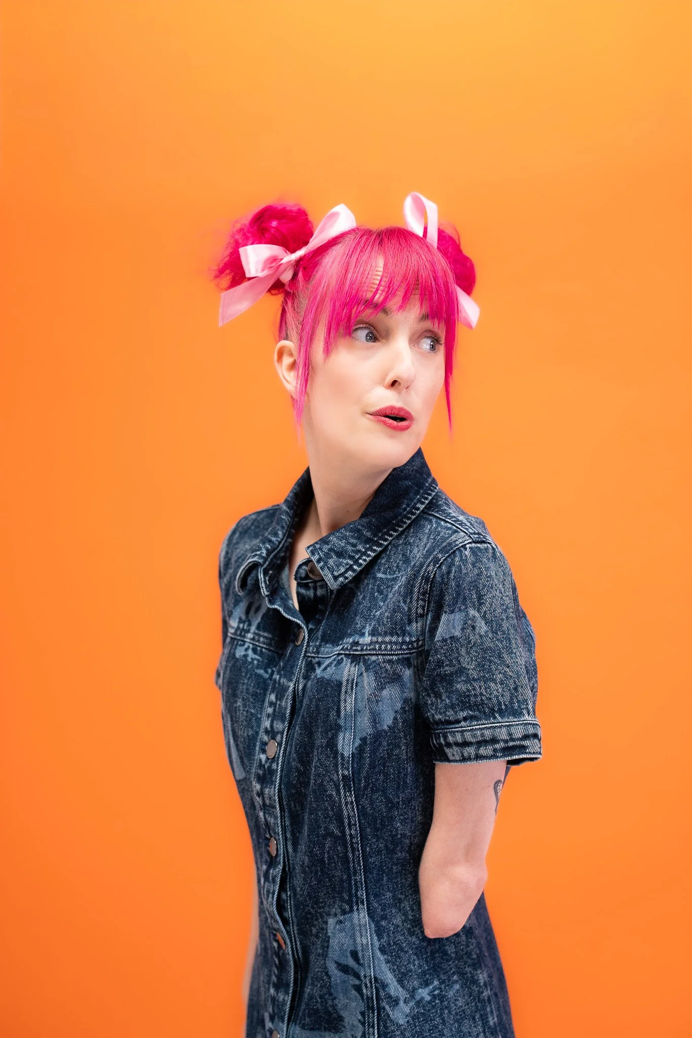 Madeleine posing in a denim dress and buns in her hair in front of an orange backdrop