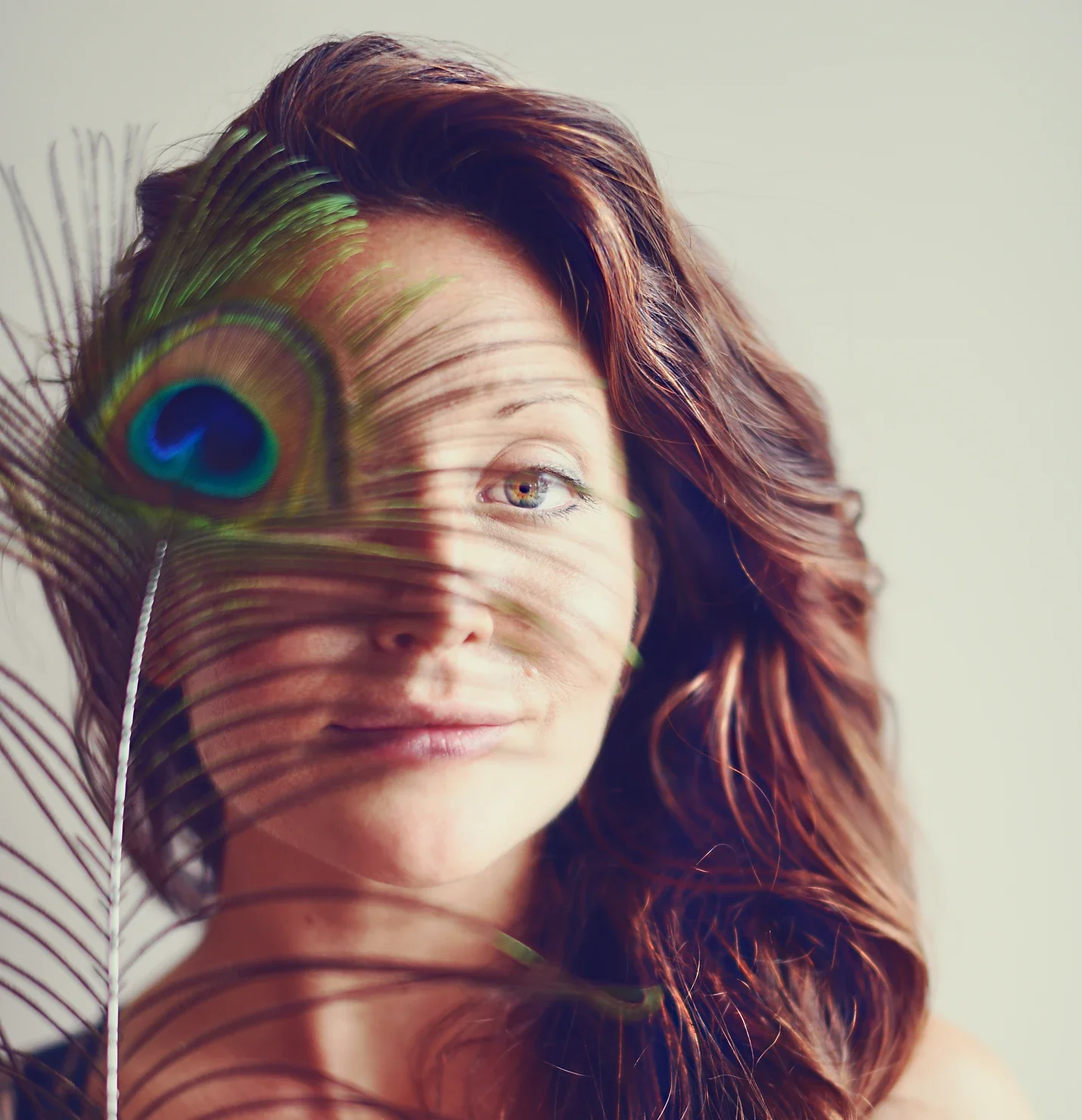 A woman with red hair holding a peacock feather in front of her face, partially covering her face and showing one eye, against a neutral background.