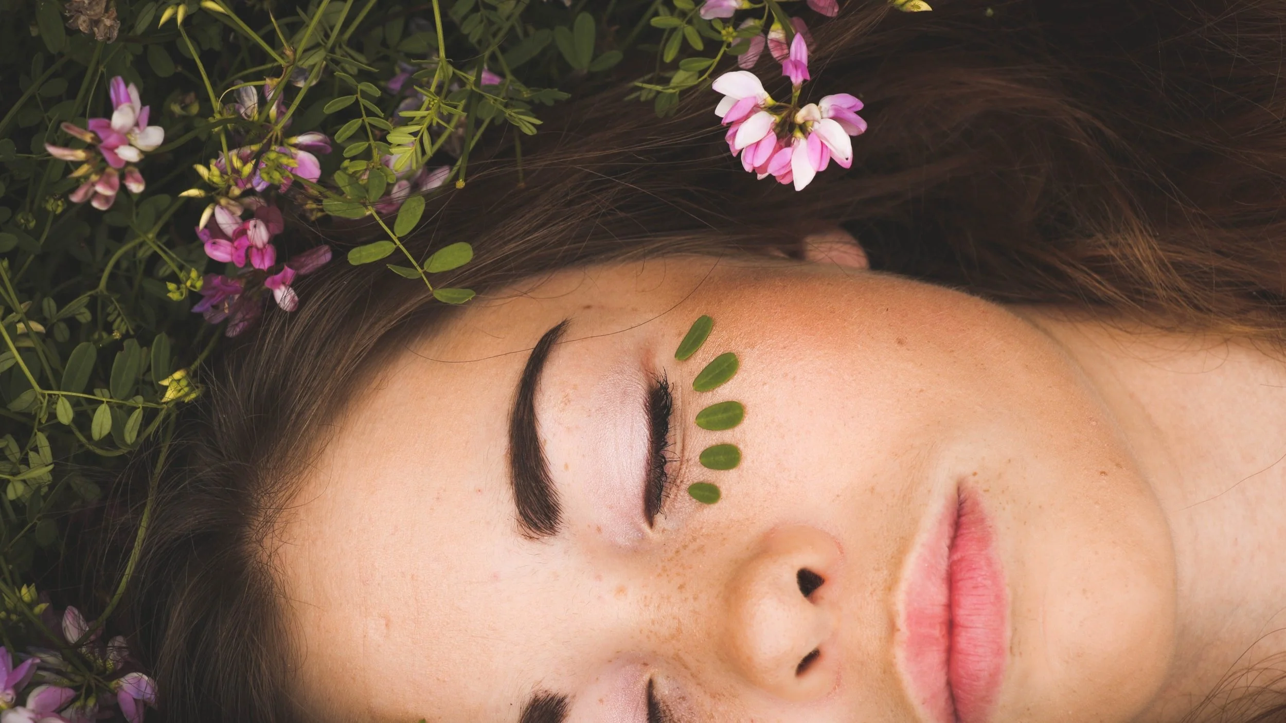 Close-up of a woman's face with closed eyes, surrounded by pink and white flowers and greenery, with green stamps arranged on her forehead.