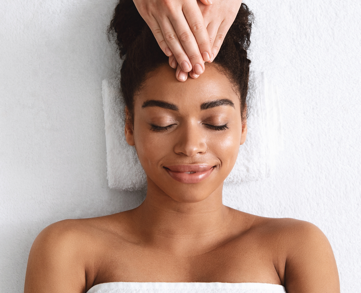 A woman receiving a facial massage while lying down with her eyes closed and a relaxed expression.