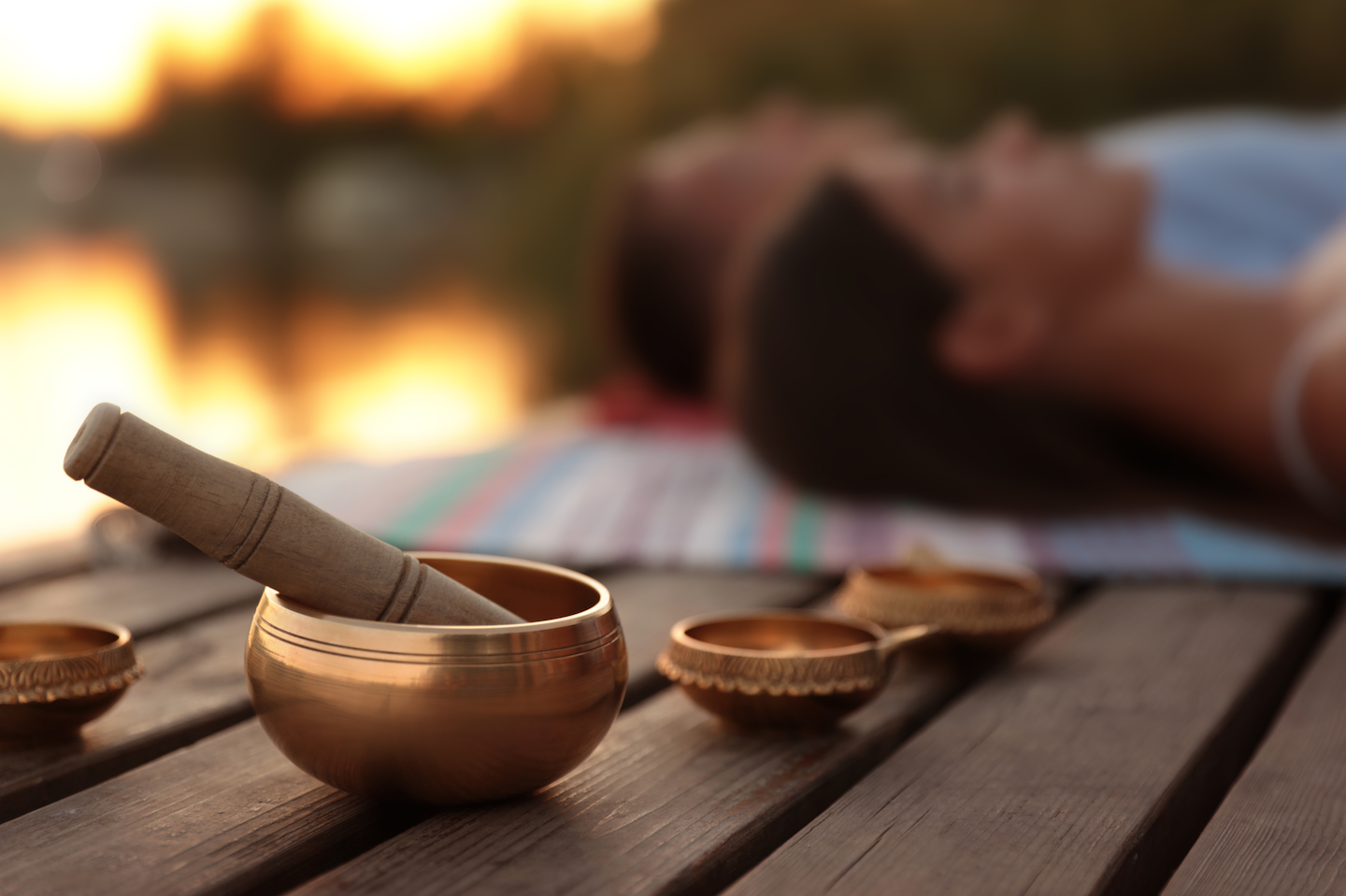 A person lying on a blanket near a fire, with a singing bowl and a wooden mallet in the foreground, outdoor setting at sunset.