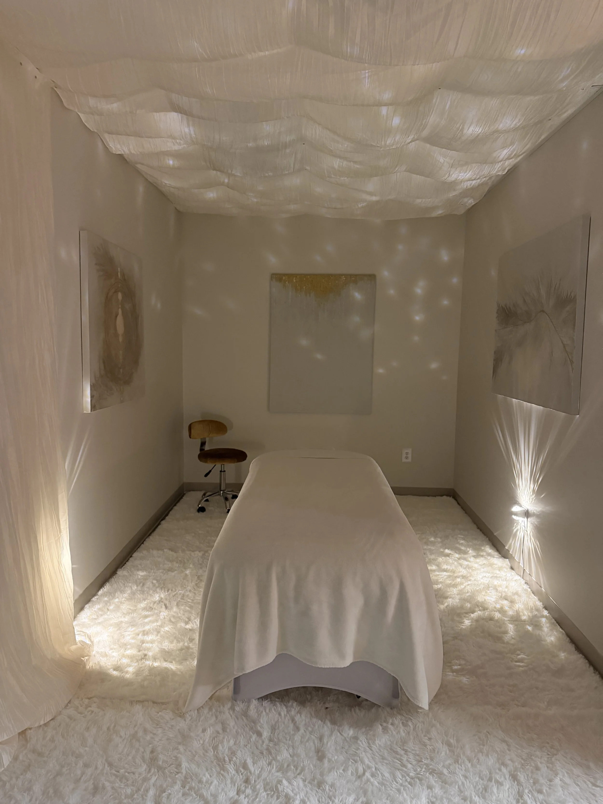 A room with soft lighting, a bed with white sheets, a fluffy white rug, two abstract wall art pieces, a mirror on the back wall, and a light fixture on the wall.