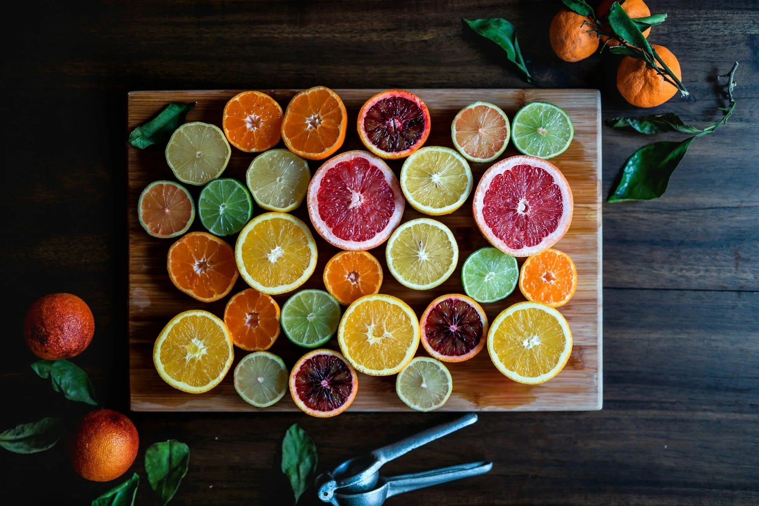 An overhead view of sliced citrus fruits on a wooden cutting board, with whole oranges and tangerines nearby and a dark wooden surface in the background.