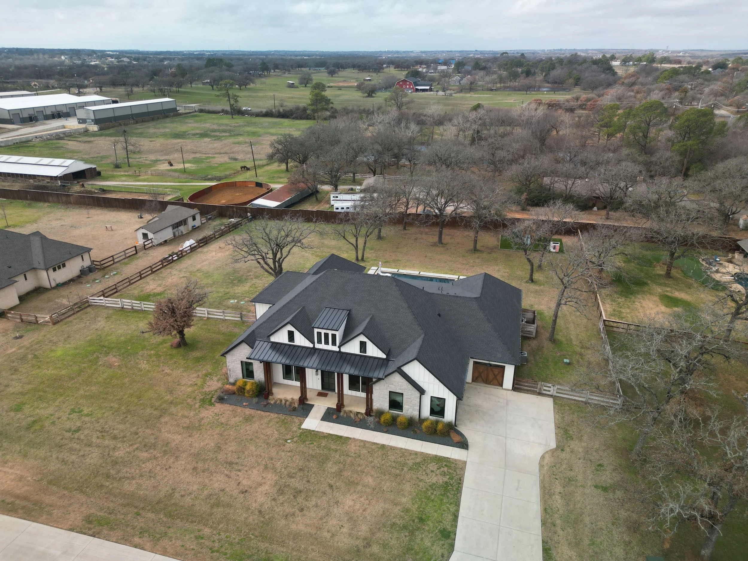 An aerial view of a suburban house with a driveway, surrounded by a large yard. The house has a modern design with black and white exterior. The yard has a lawn and some bushes in front of the house. There are trees and a fence around the backyard, a