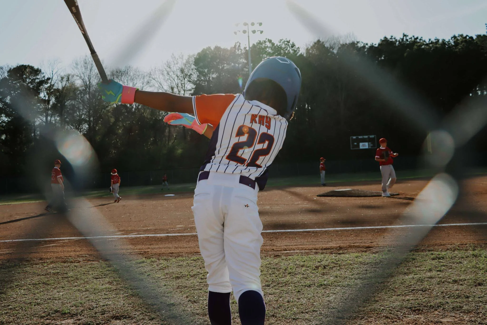 A young baseball player wearing a blue helmet, striped jersey with the number 22, and gloves, swinging a bat during a game on a sunny day. Other players are visible on the field.