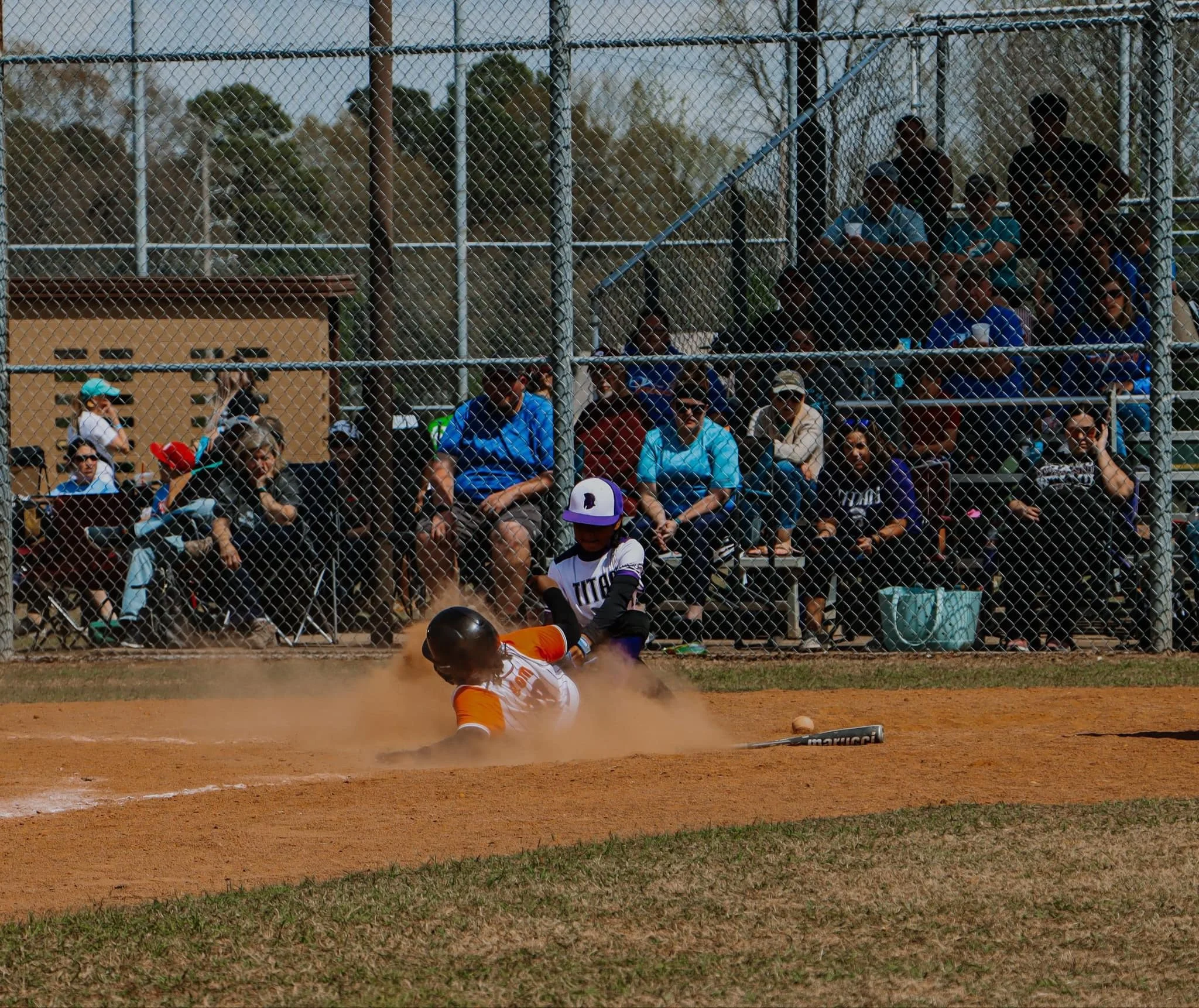A baseball player in an orange uniform sliding into home plate during a game, with spectators watching from behind a chain-link fence.