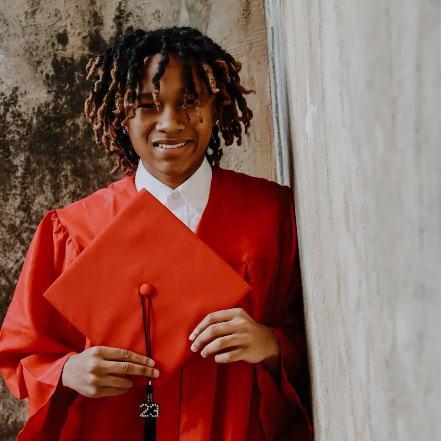 A young man in a red graduation gown and cap holding a tassel with the number 23, smiling, standing against a beige wall.