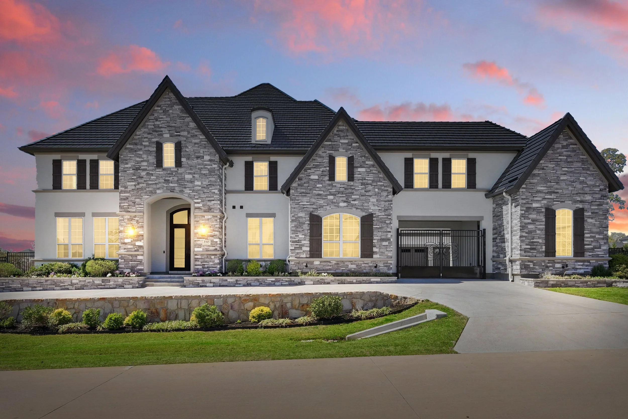Front view of a two-story house with stone and stucco exterior, multiple gable roofs, illuminated windows, a black front gate, and a landscaped lawn during twilight.