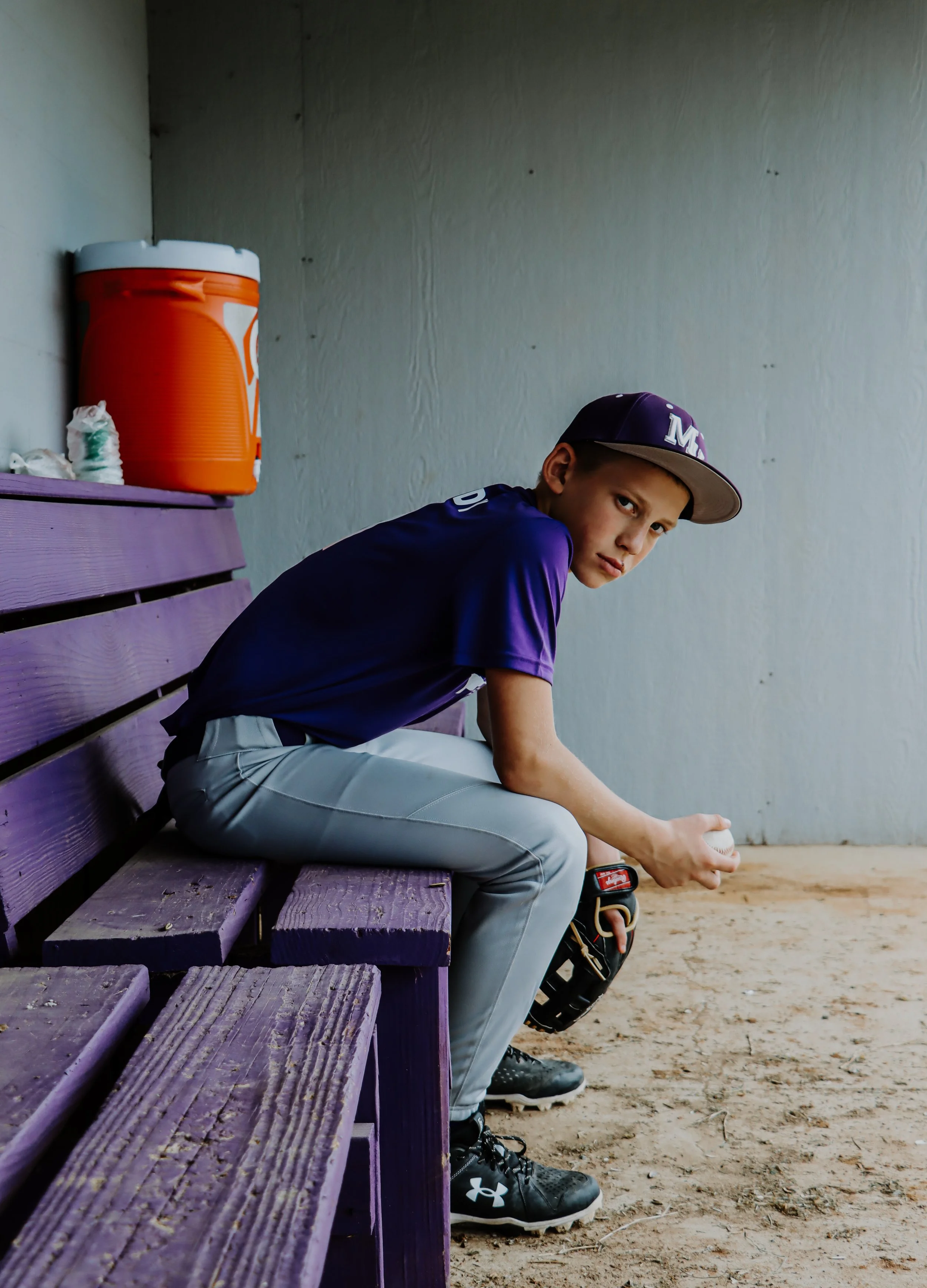 A young baseball player sitting on a purple bench in a dugout, holding a baseball, wearing a purple cap and uniform, with a glove and a baseball cleat on the ground next to him.