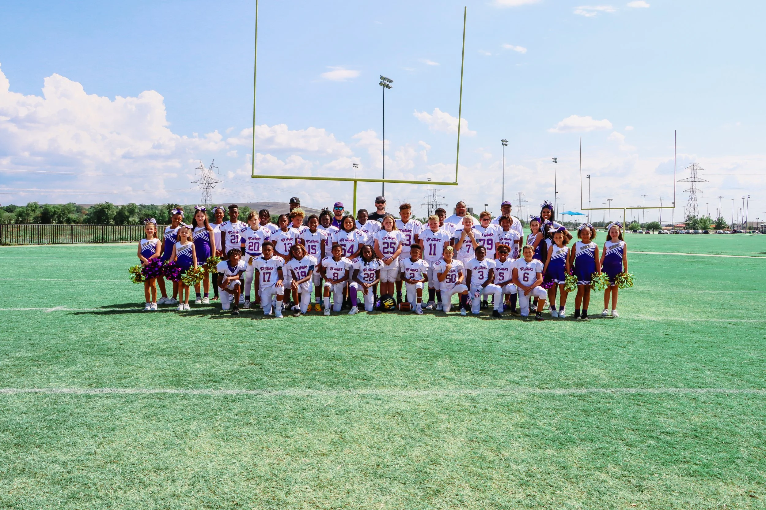 Group photo of a youth football team and cheerleaders on a grassy field with goalposts, cheerleaders holding pom-poms, and coaches behind the players, under a partly cloudy sky.