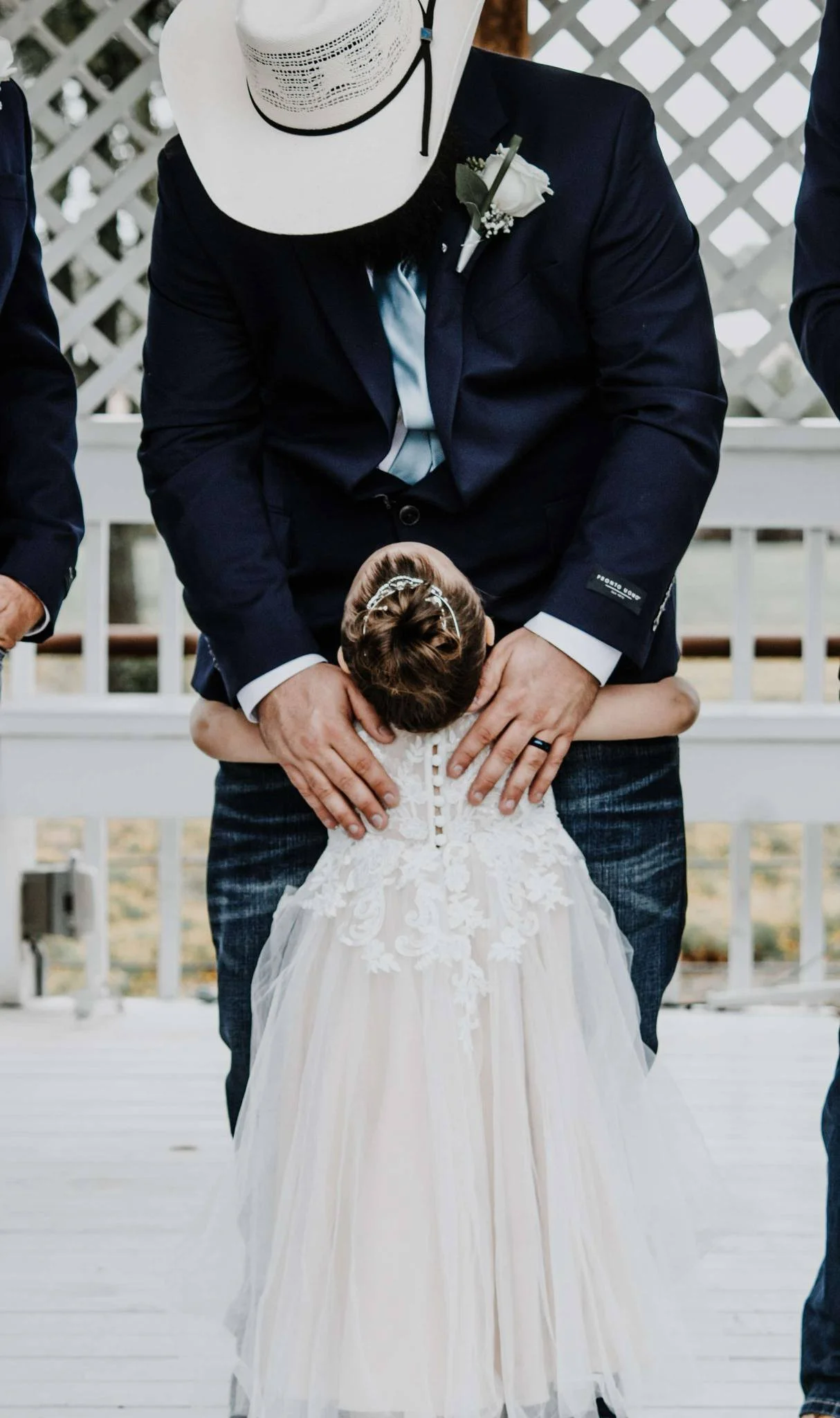 A man and a young girl, likely at a wedding or special event. The man is wearing a dark suit with a boutonnière and a light blue tie, and the girl is dressed in a white lace dress with a floral headband. The man is bowing his head and the girl is hug
