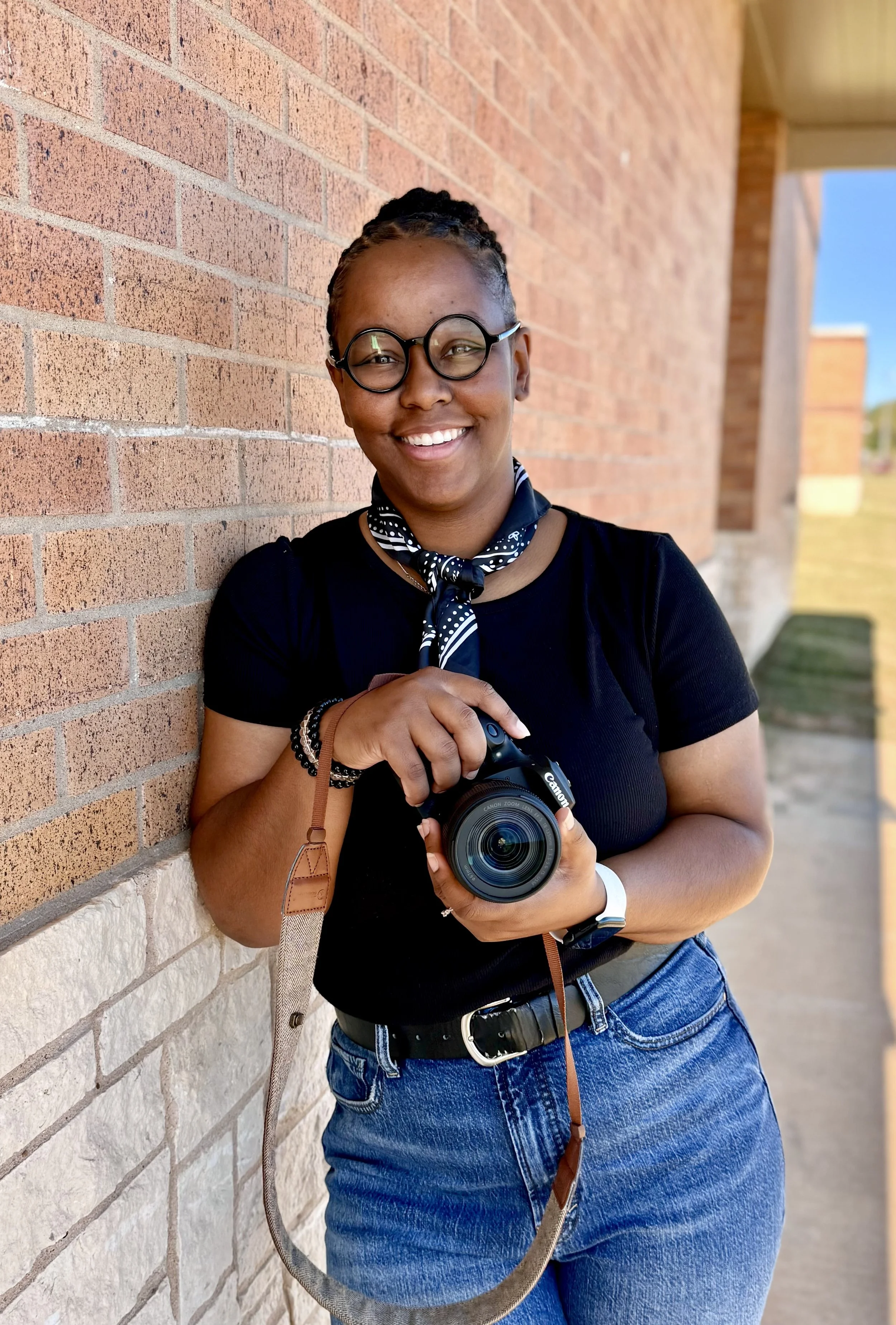 Smiling woman with glasses, polka dot scarf, and camera standing against brick wall outside.