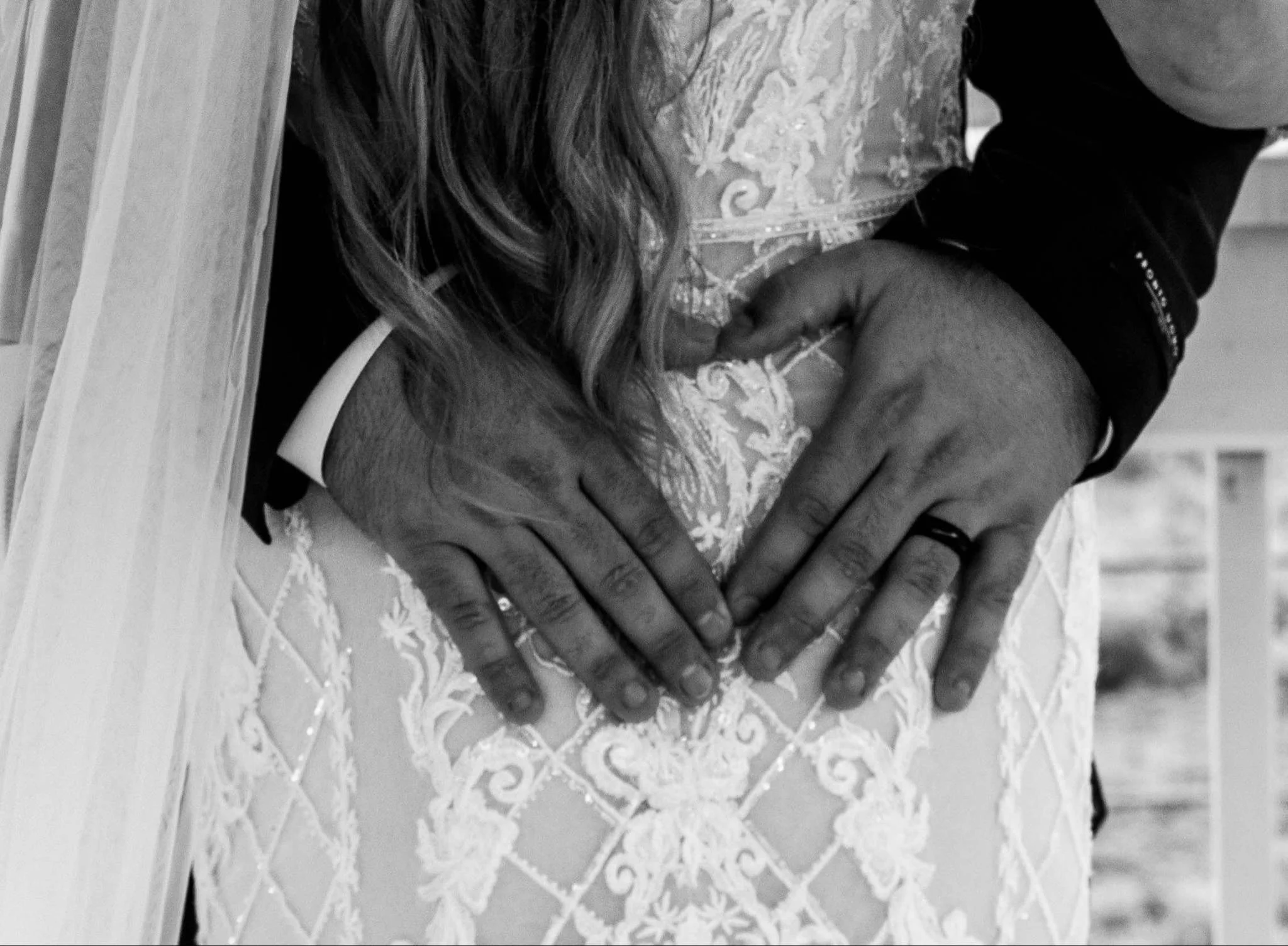 Close-up of a couple's hands holding on top of a lace wedding dress, with one person wearing a wedding ring.