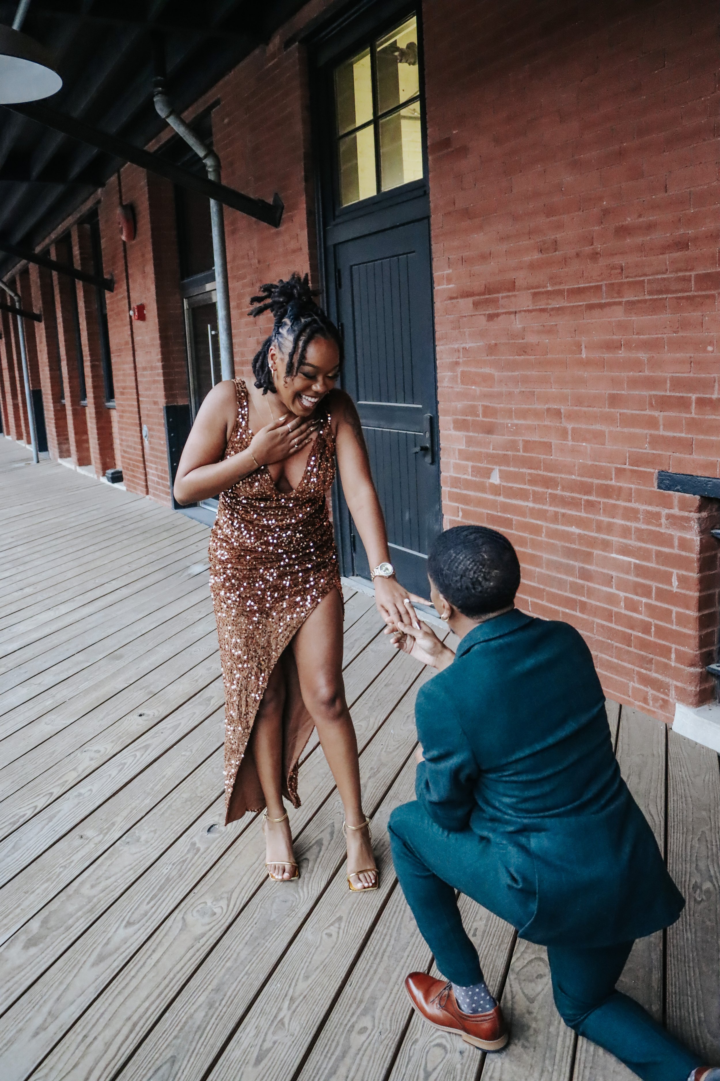 A woman in a sparkly dress is surprised and laughing as a man in a suit proposes marriage to her with a ring on an outdoor wooden deck.