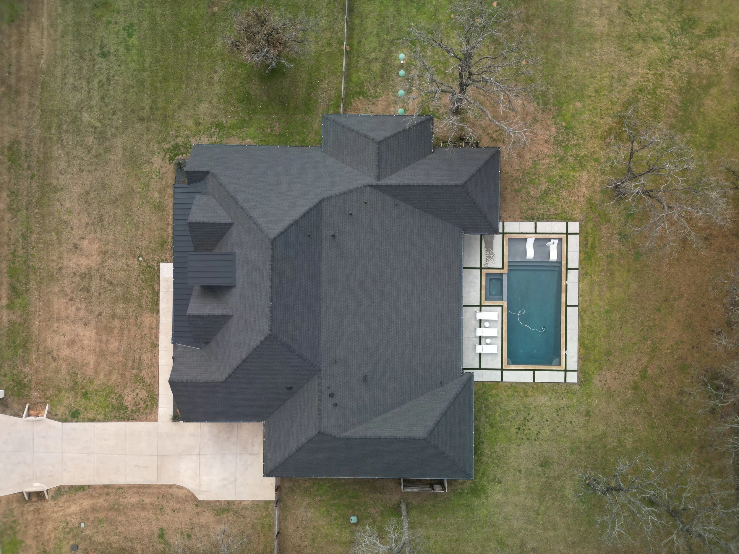 An aerial view of a house with a dark gray roof, a backyard with a swimming pool, and a grassy yard with some trees and a concrete walkway.