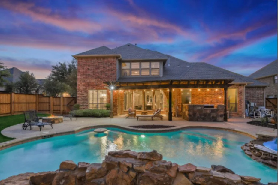 A backyard with a swimming pool, patio furniture, and a two-story brick house at sunset.