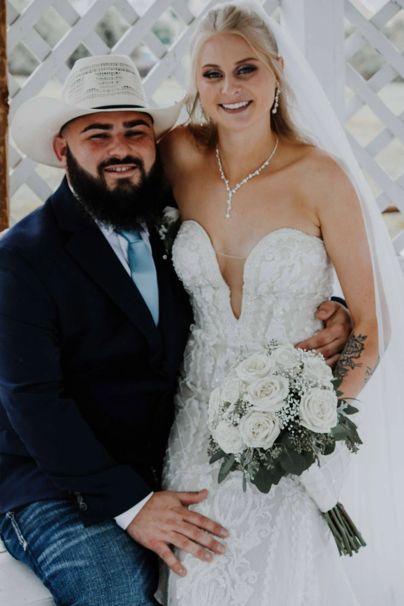 A newlywed couple sitting together, with the bride holding a bouquet of white roses, both dressed in wedding attire, smiling, in front of a lattice backdrop.