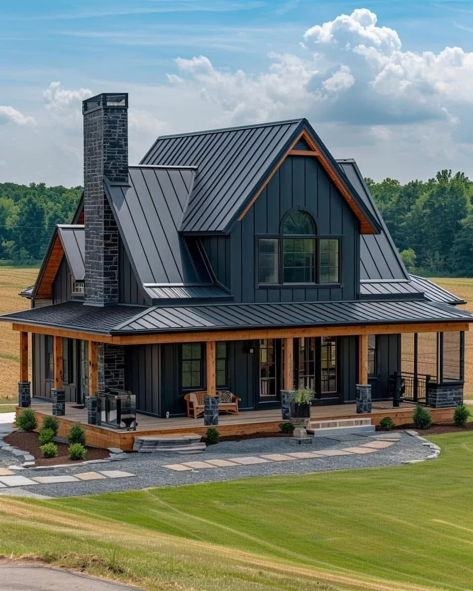 A modern two-story house with black metal roofing, dark blue siding, and a spacious wooden porch, situated in a rural area with green fields and trees in the background under a partly cloudy sky.