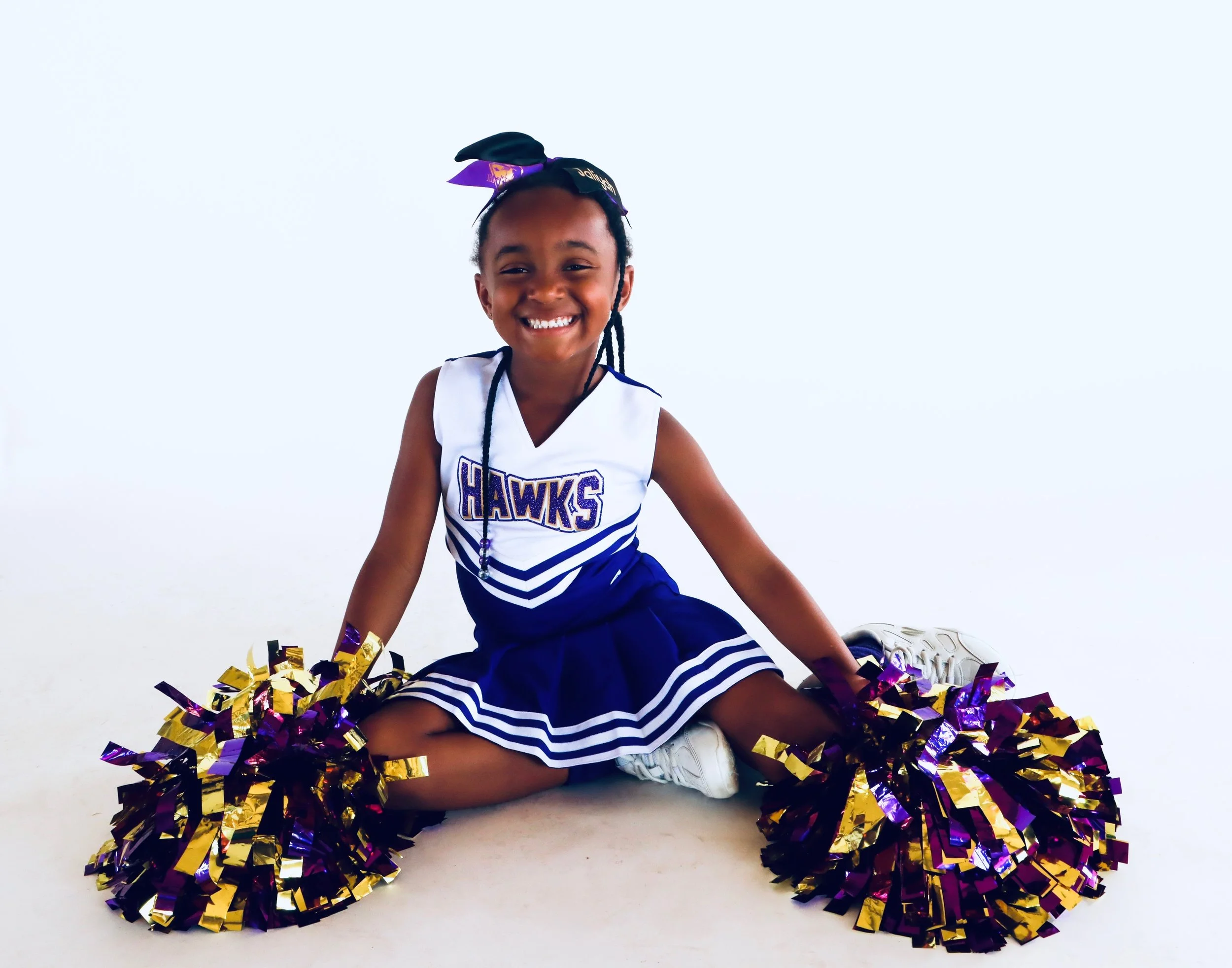 Smiling young girl dressed as a cheerleader with a purple and gold bow in her hair, sitting on the floor amidst purple and gold pom-poms, wearing a white and purple cheerleading uniform with 