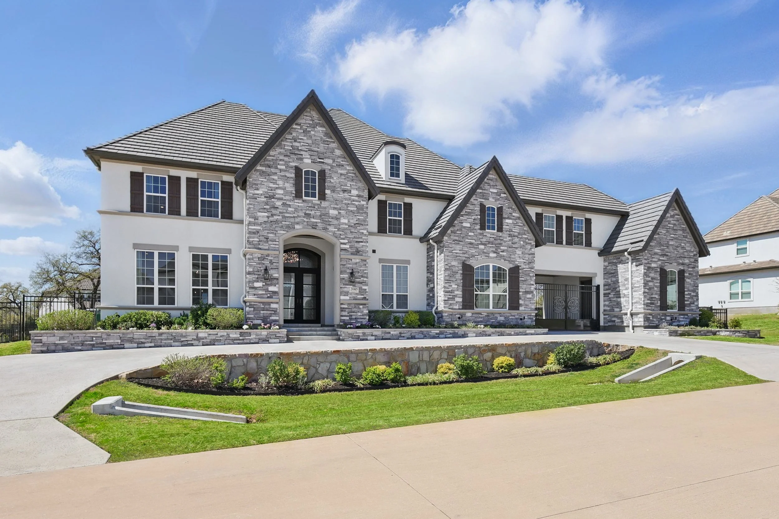 Front view of a modern two-story house with white and gray brick exterior, multiple windows, and a front yard with a curved driveway and landscaped garden.