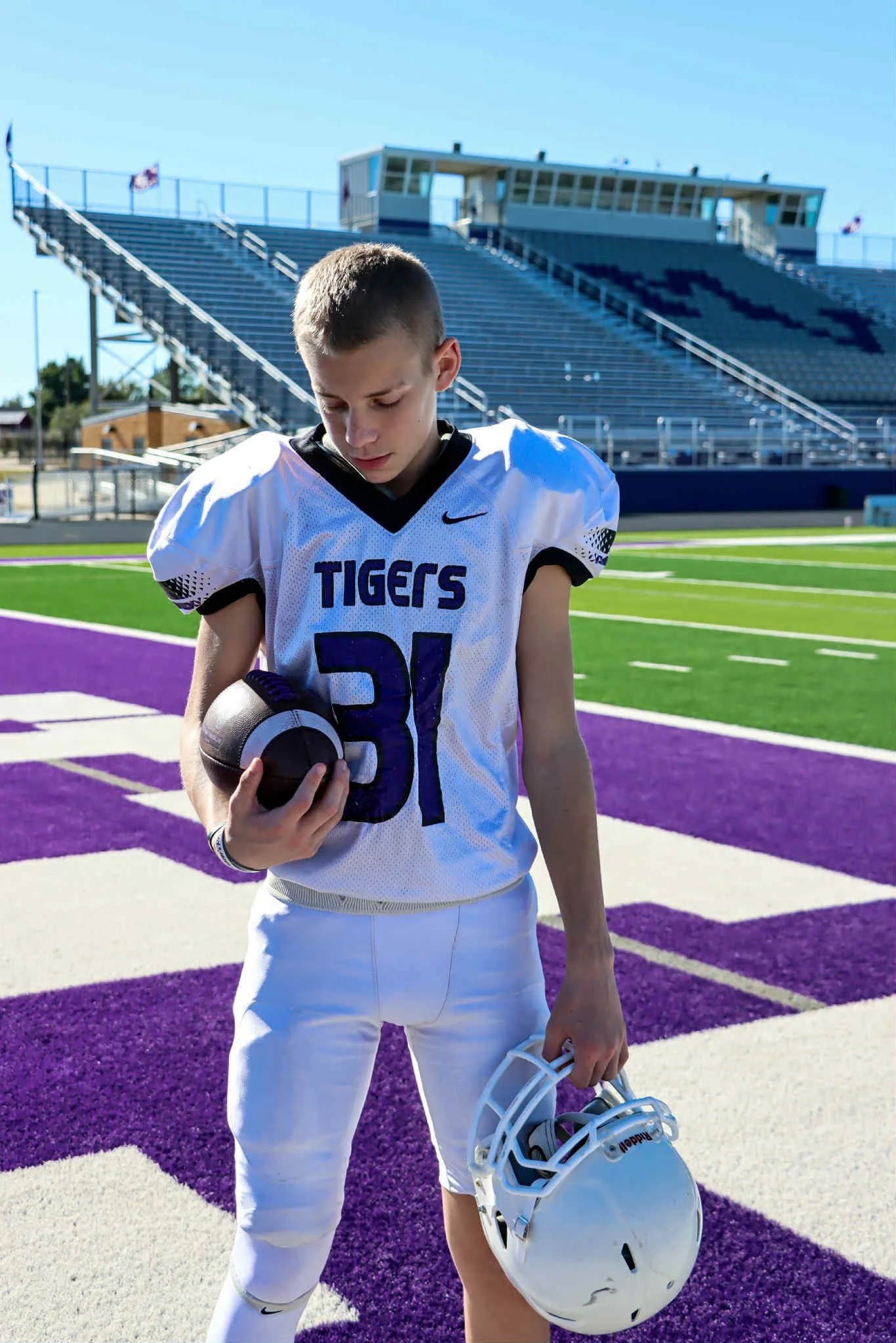 A young football player in a white uniform holding a helmet and a football on a colorful football field with purple and white markings and empty stadium bleachers in the background.