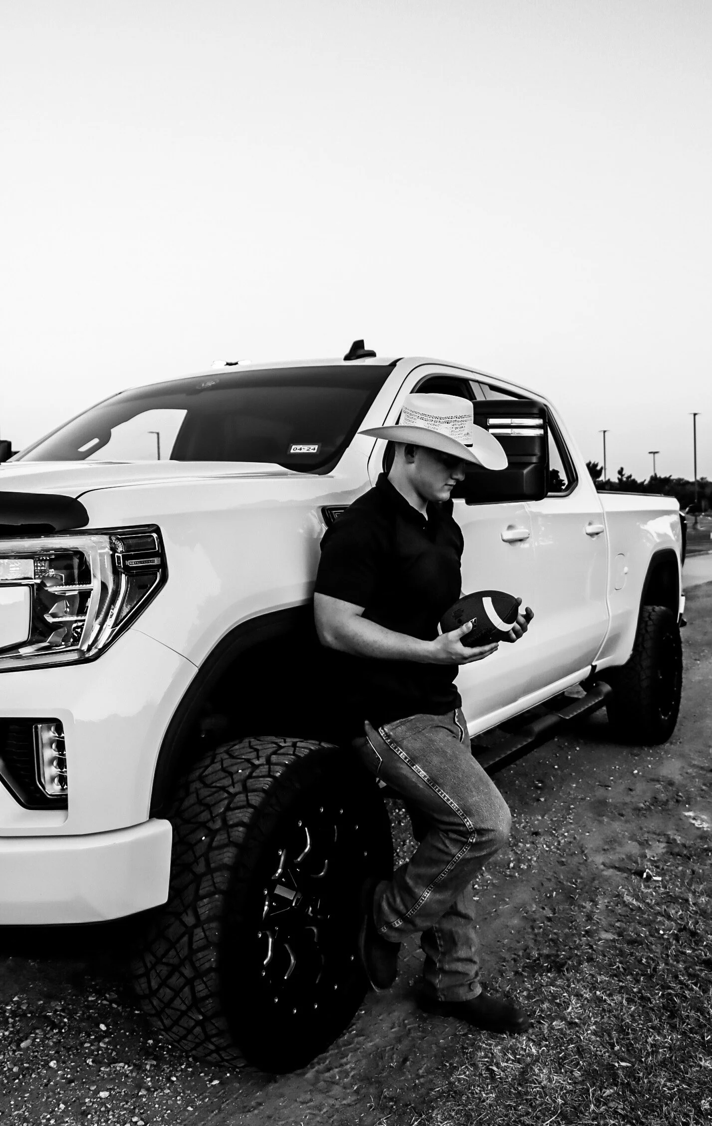 A person wearing a cowboy hat and casual clothes leaning against a large white pickup truck holding a football in their hands, in an outdoor parking lot.