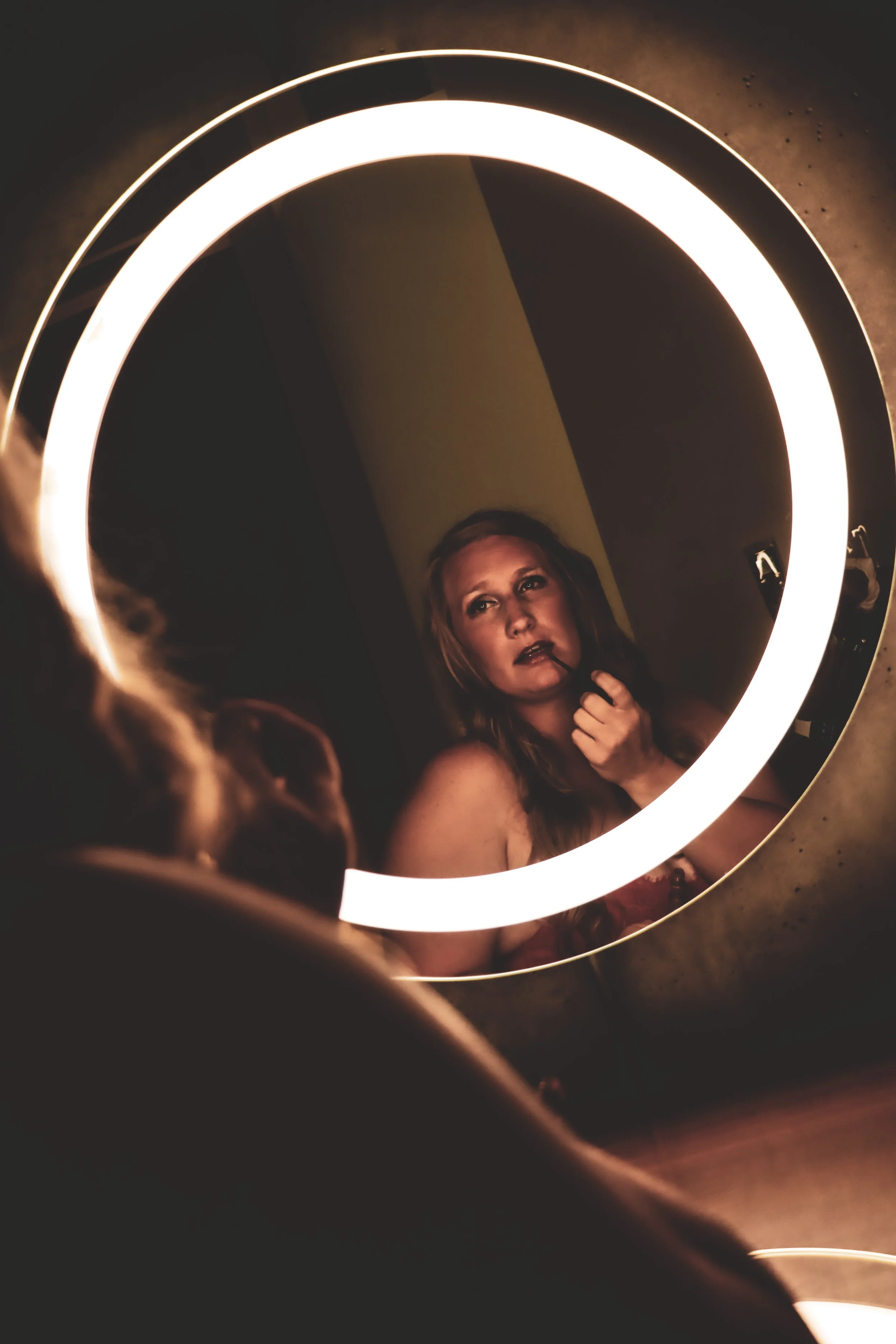 A woman with red hair applying lipstick while looking into a round illuminated mirror in a dimly lit room.