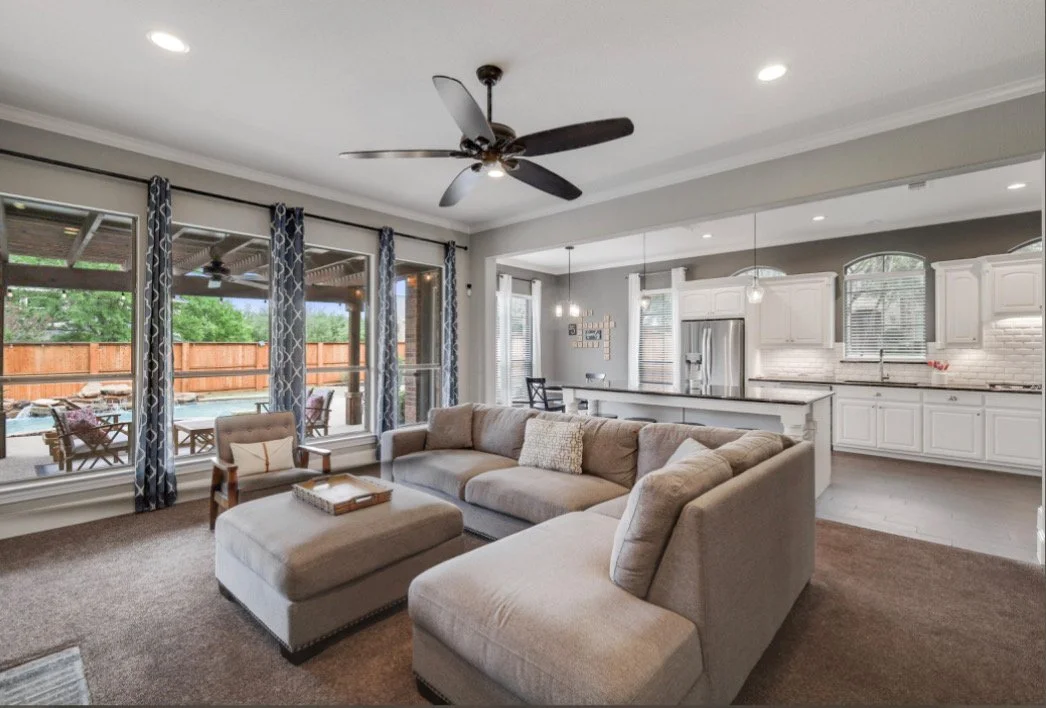 Living room with beige sectional sofa, ottoman, large windows with patterned curtains, view of outdoor patio, white kitchen with stainless steel appliances, pendant lights, and bar stools in an open floor plan.