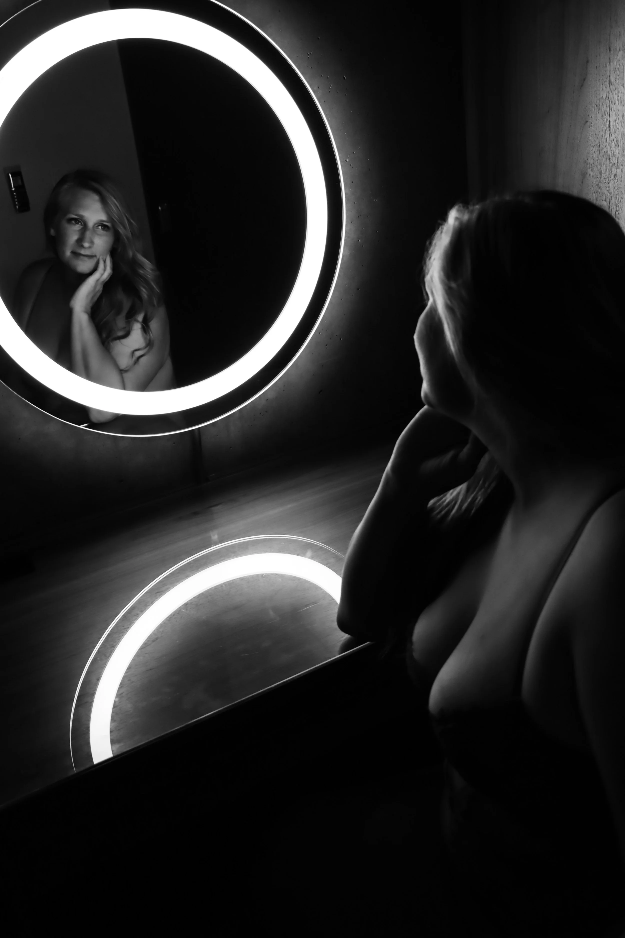 A woman with long hair looks into a circular mirror with neon lighting, her reflection visible, while another woman sits nearby, resting her chin on her hand, in a dimly lit room.