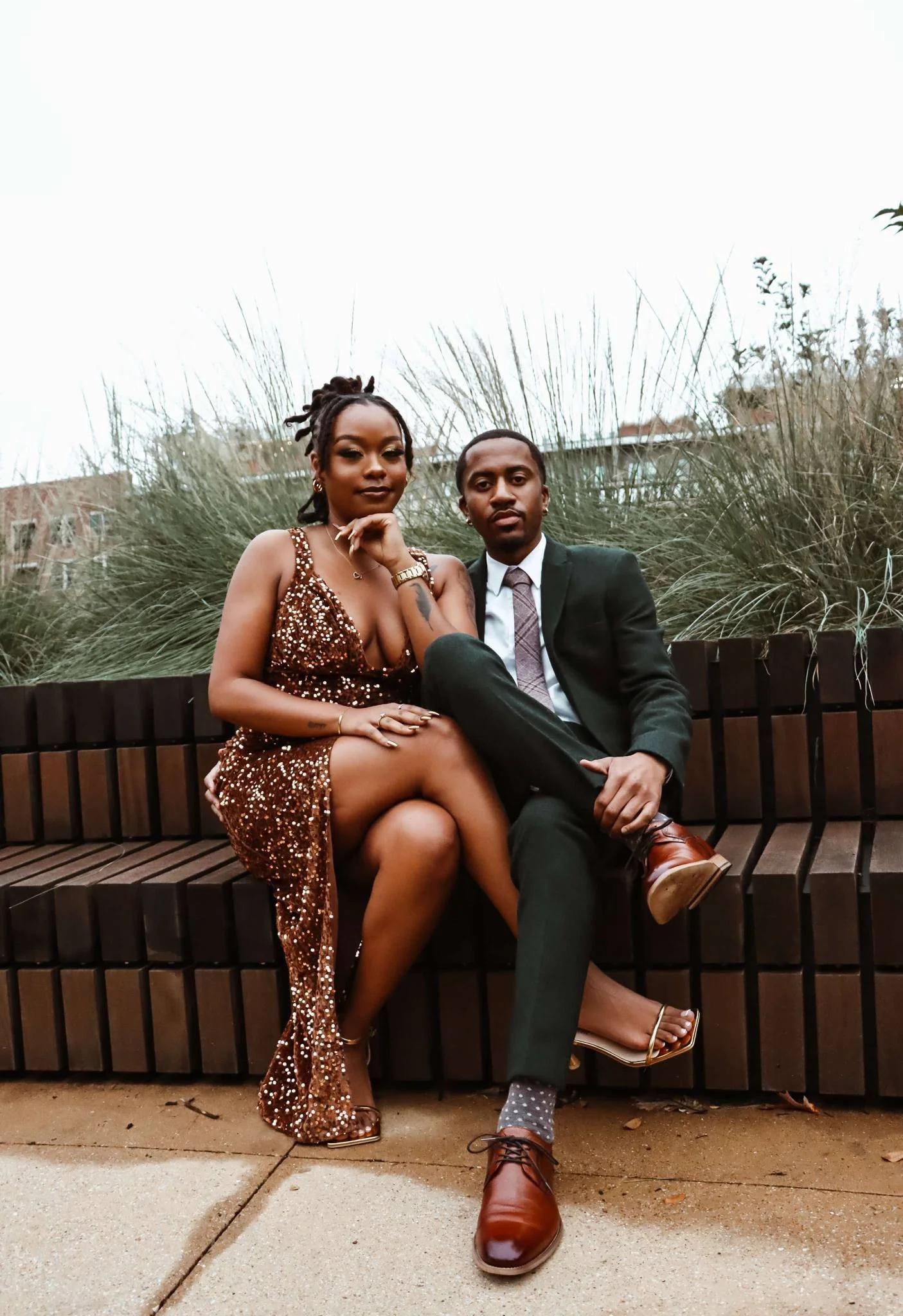A man and a woman sitting on a wooden bench outdoors. The woman is dressed in a sparkly, bronze-colored dress with a deep V neckline, and the man is wearing a dark suit with a light-colored shirt and patterned tie. They are sitting close together, lo